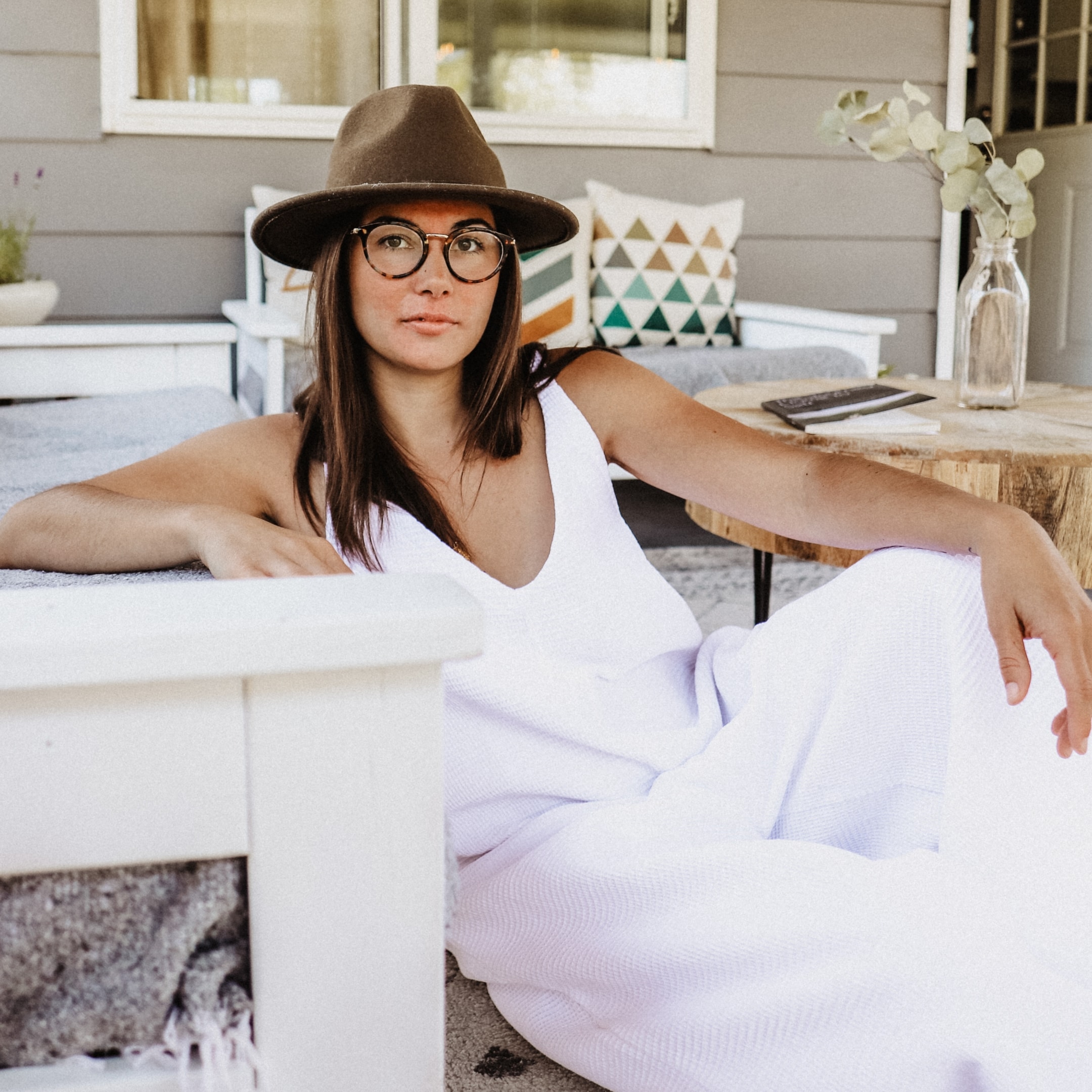 Woman wearing glasses and a wide-brimmed hat sitting outdoors on a porch with cushions and a table.