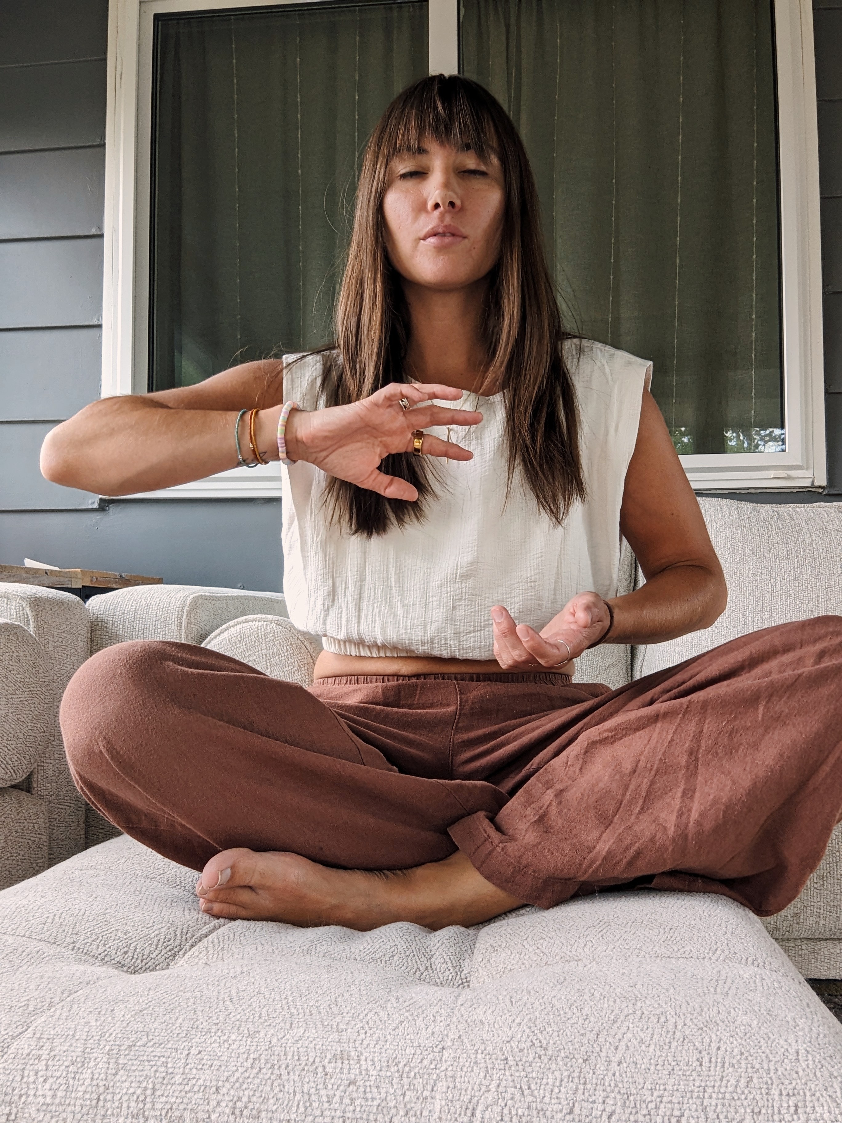 Woman sitting cross-legged on a couch with eyes closed, practicing meditation or mindfulness, in front of a window with curtains.