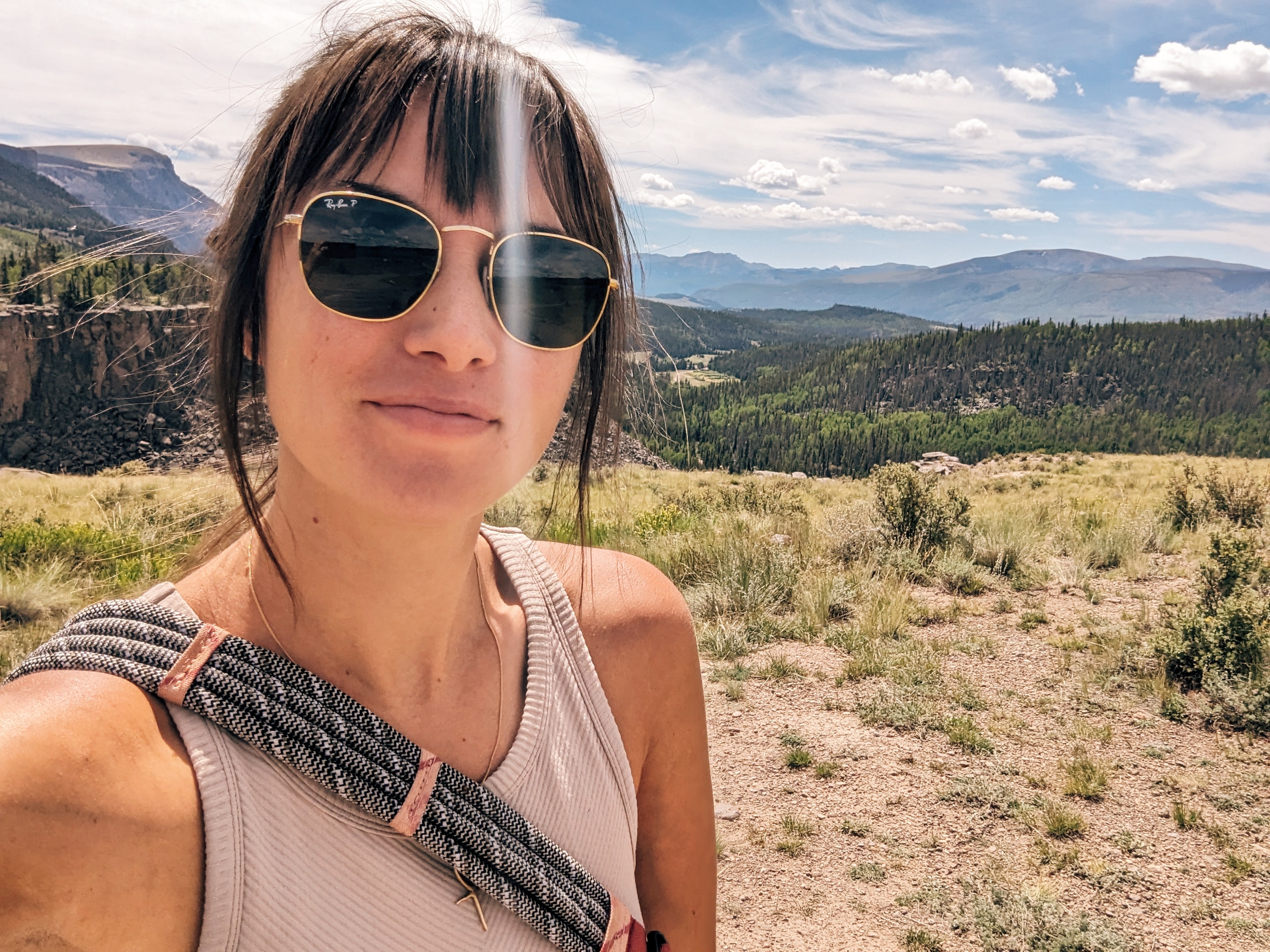 Woman with sunglasses outdoors in a landscape with mountains and sky, wearing a sleeveless top and carrying a bag.