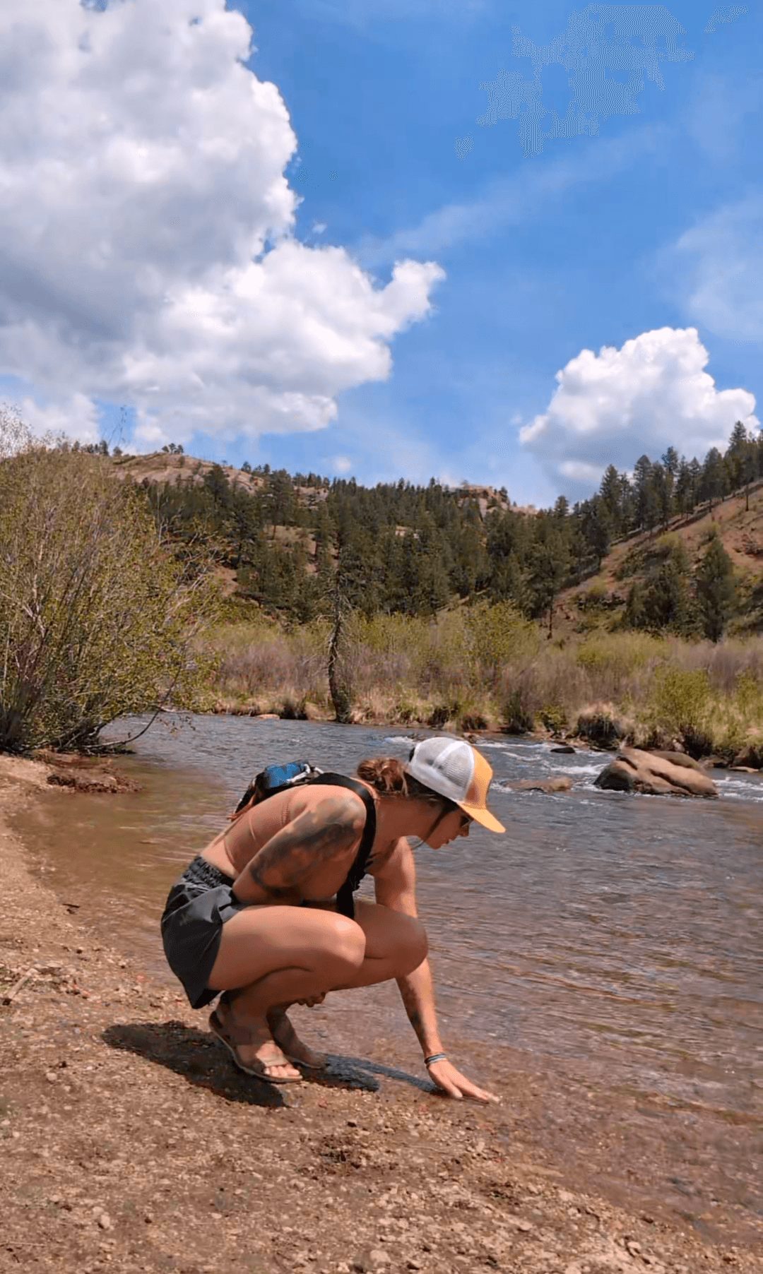 Person crouching by a riverbank wearing a hat and swimsuit, with trees and hills in the background under a partly cloudy sky.