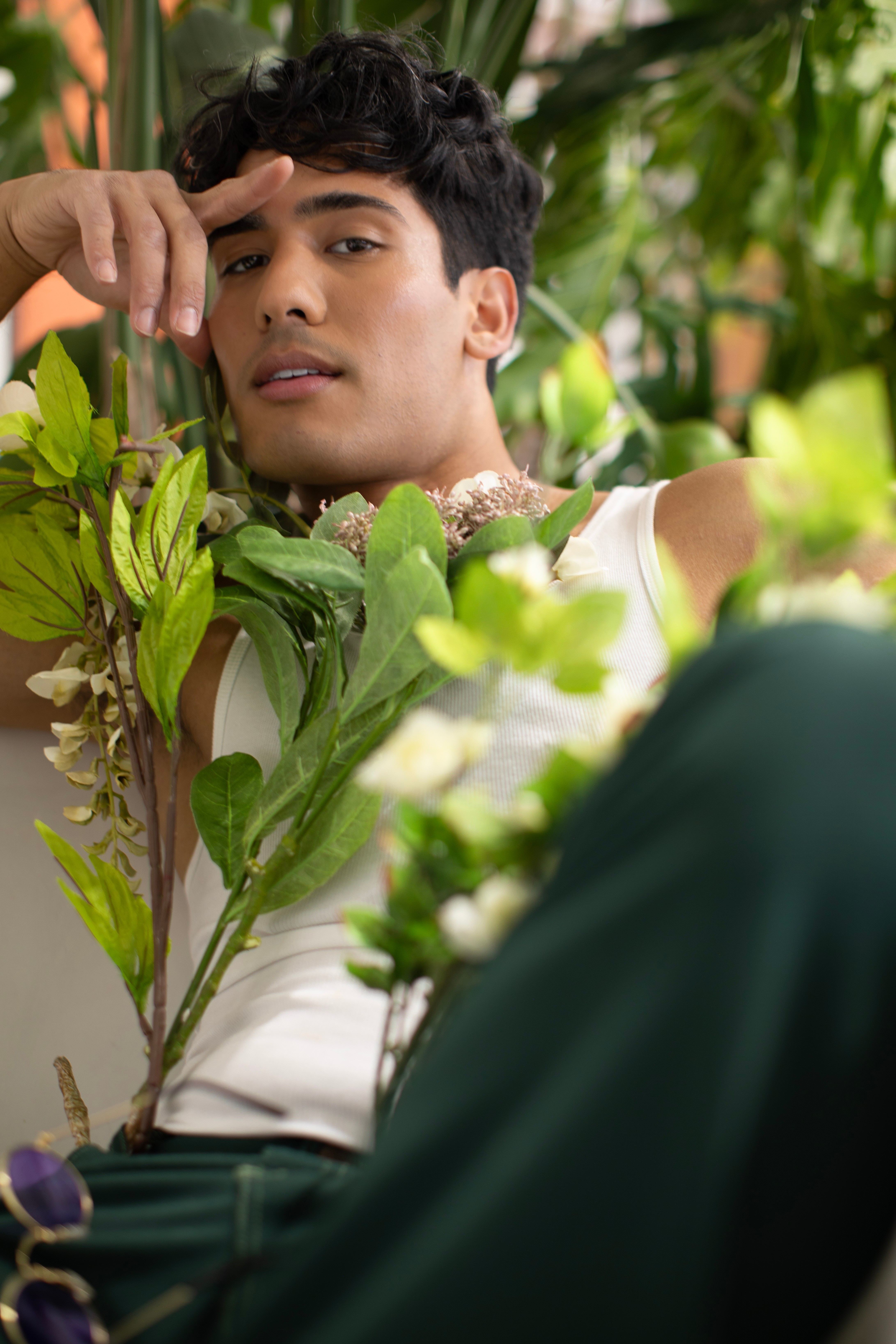 Young man with dark hair lying among green plants, looking at the camera, hand near forehead, surrounded by foliage.
