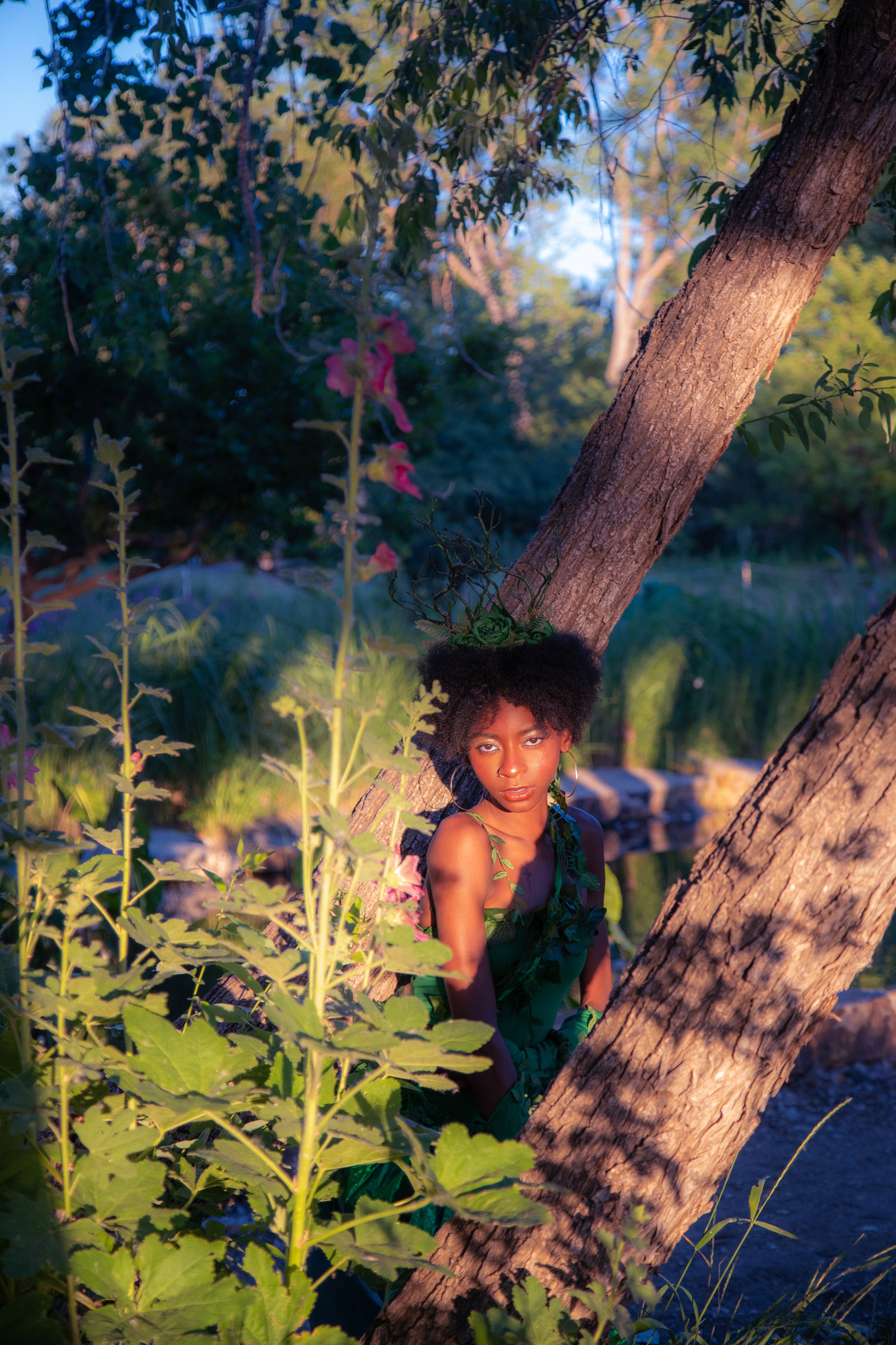 Young girl with curly hair standing among tall plants near a tree by a pond, sunlight filtering through leaves.