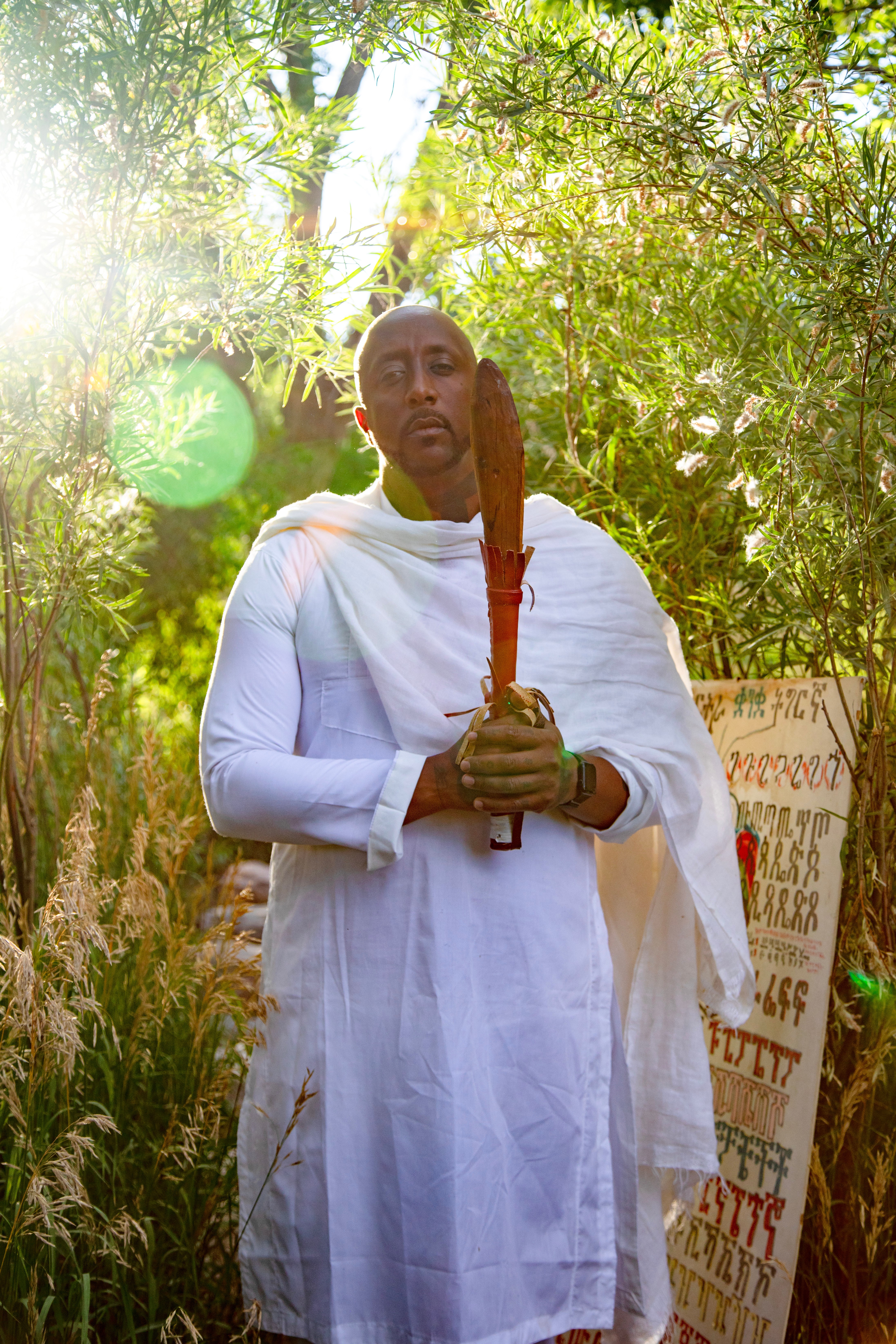 Person dressed in white holding a wooden staff outdoors with greenery and sunlight in the background.