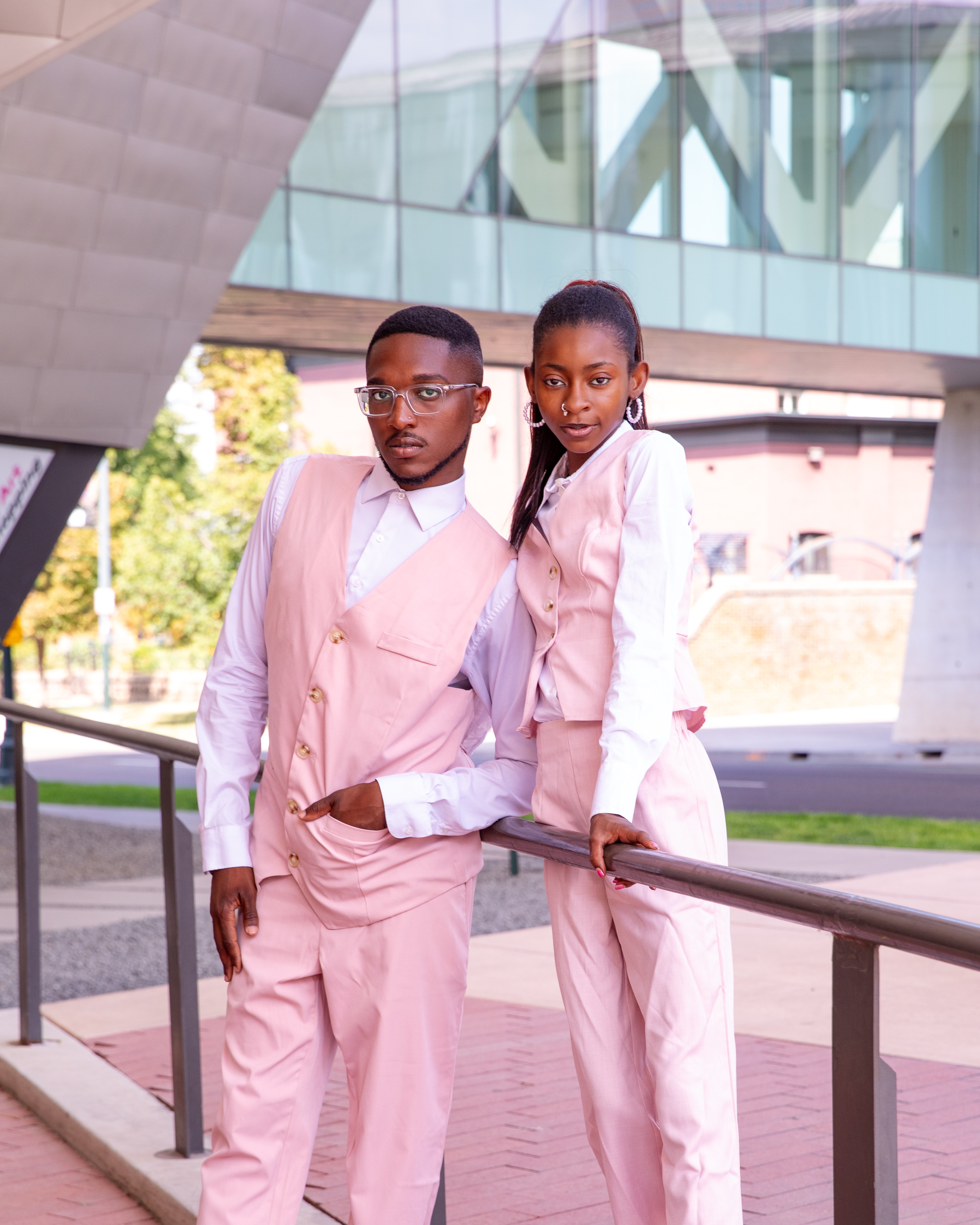 Two people in pink suits standing outdoors near a railing, with a modern building in the background.