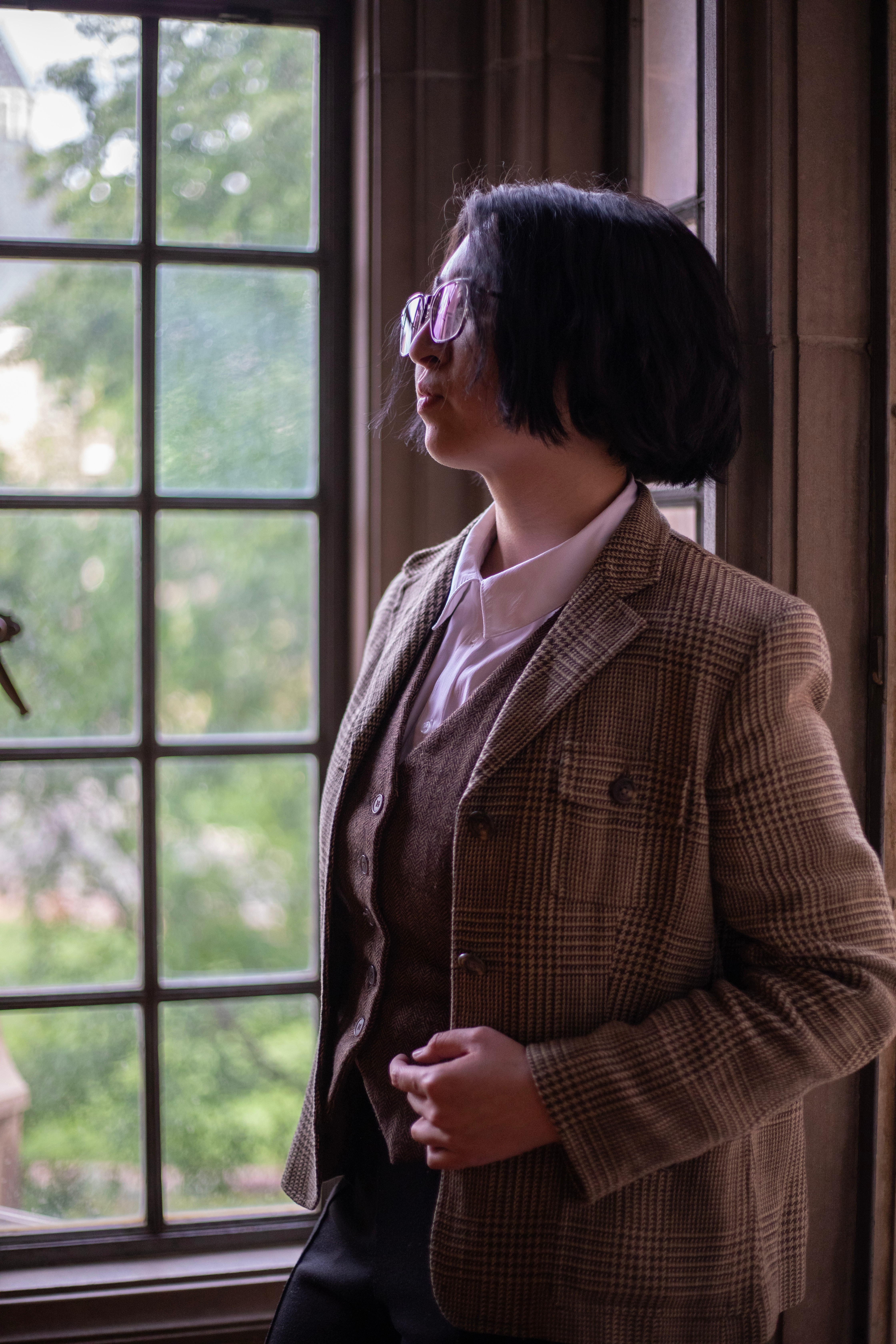 Person with short dark hair wearing a brown checkered blazer standing by a window, looking outside.