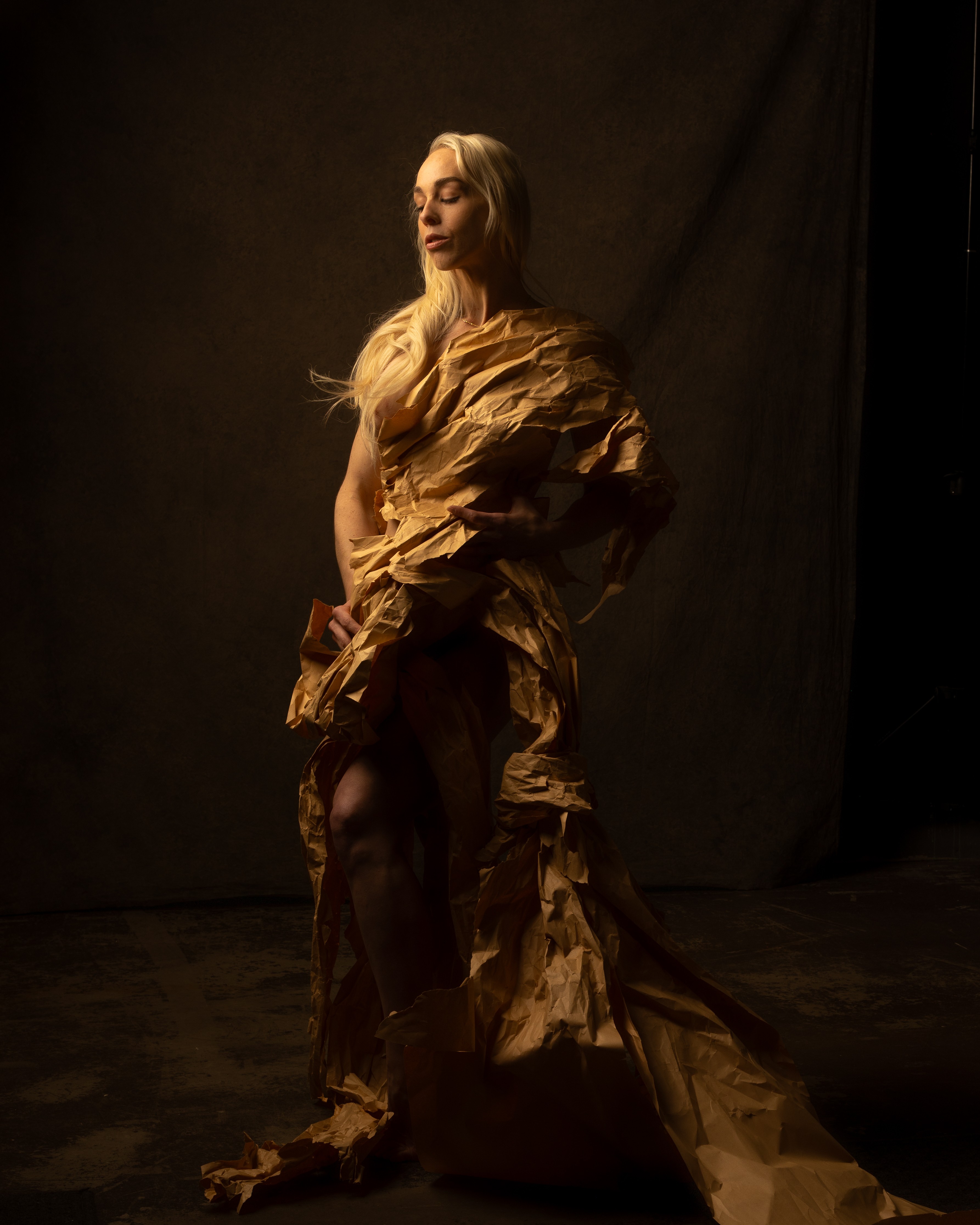 A woman with long blonde hair stands in a dark setting, wearing a flowing, textured brown dress with dramatic folds.