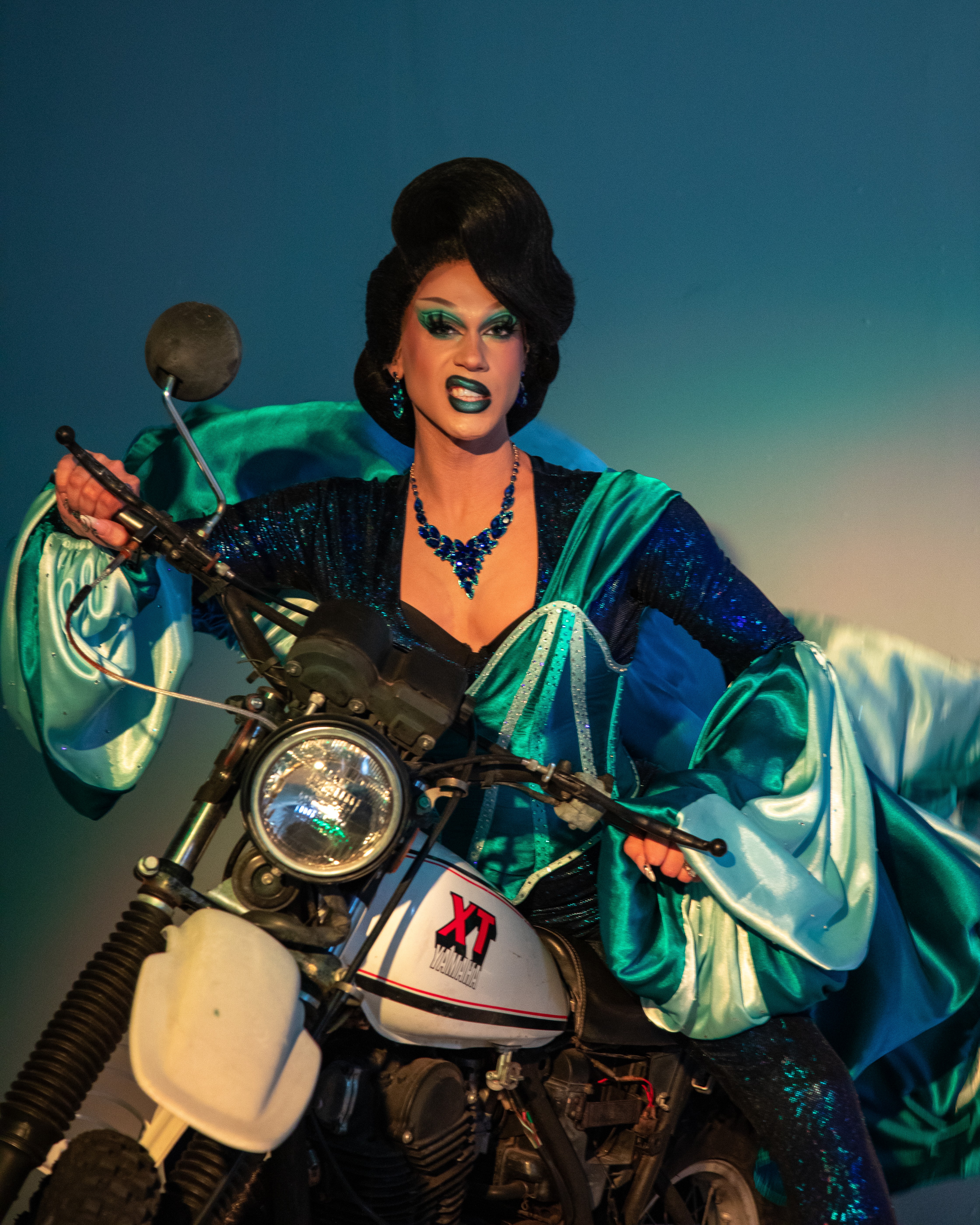 Person with dark hair and makeup riding a motorcycle, wearing a shiny blue and green outfit, against a gradient background.