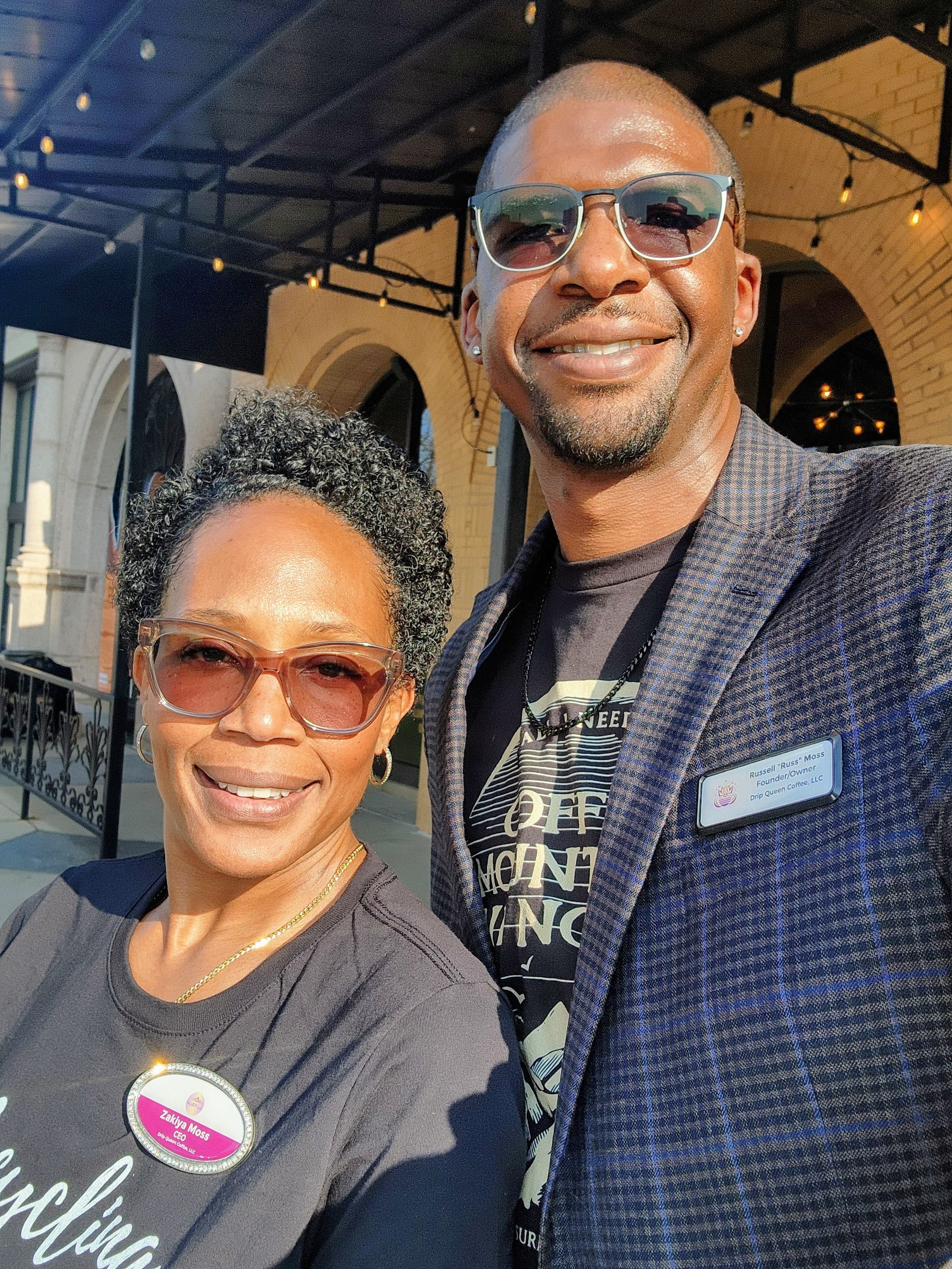 Two smiling people wearing sunglasses outdoors, with a building and string lights in the background.