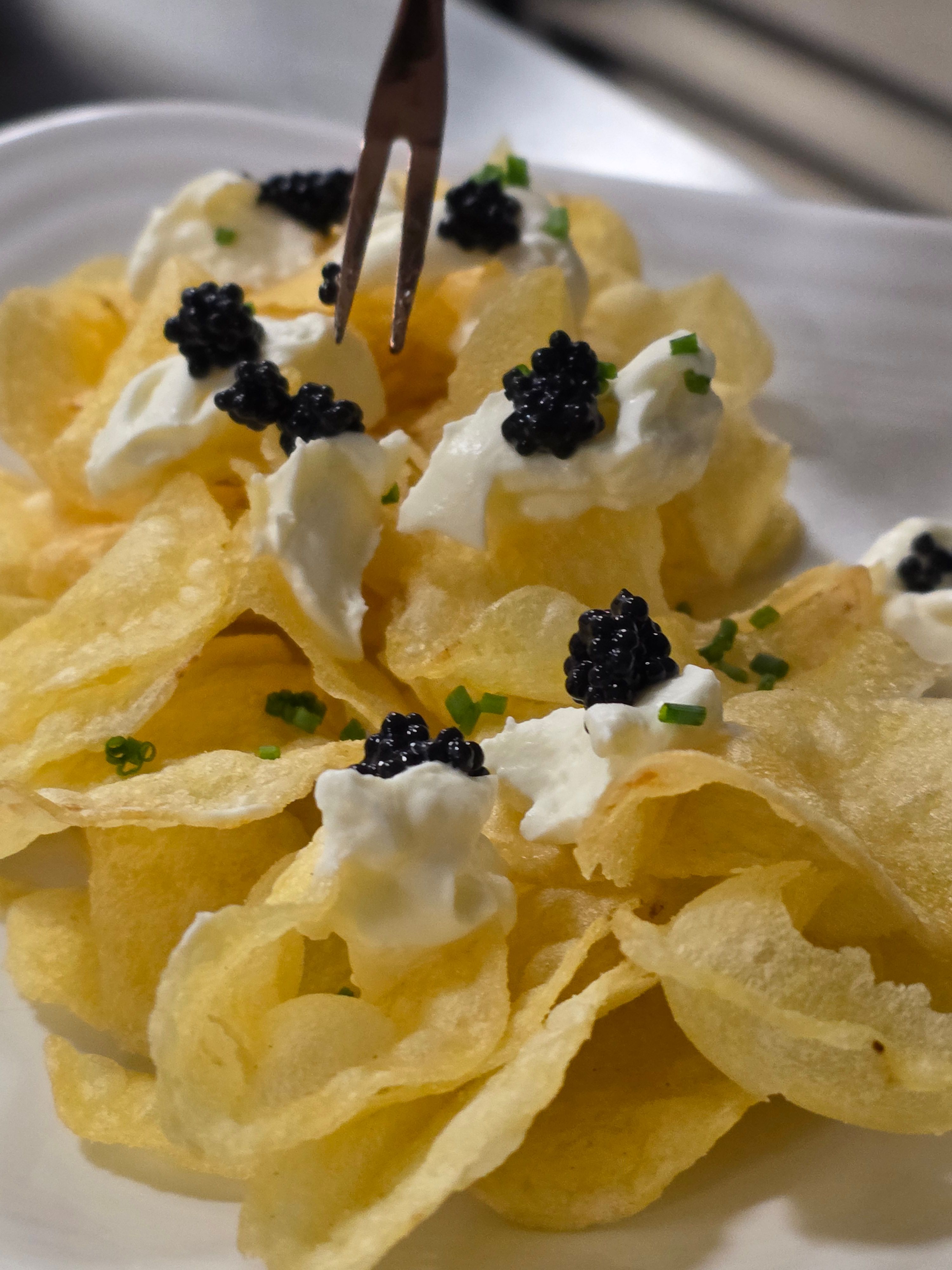 Potato chips topped with dollops of cream, black caviar, and chopped green herbs on a white plate.