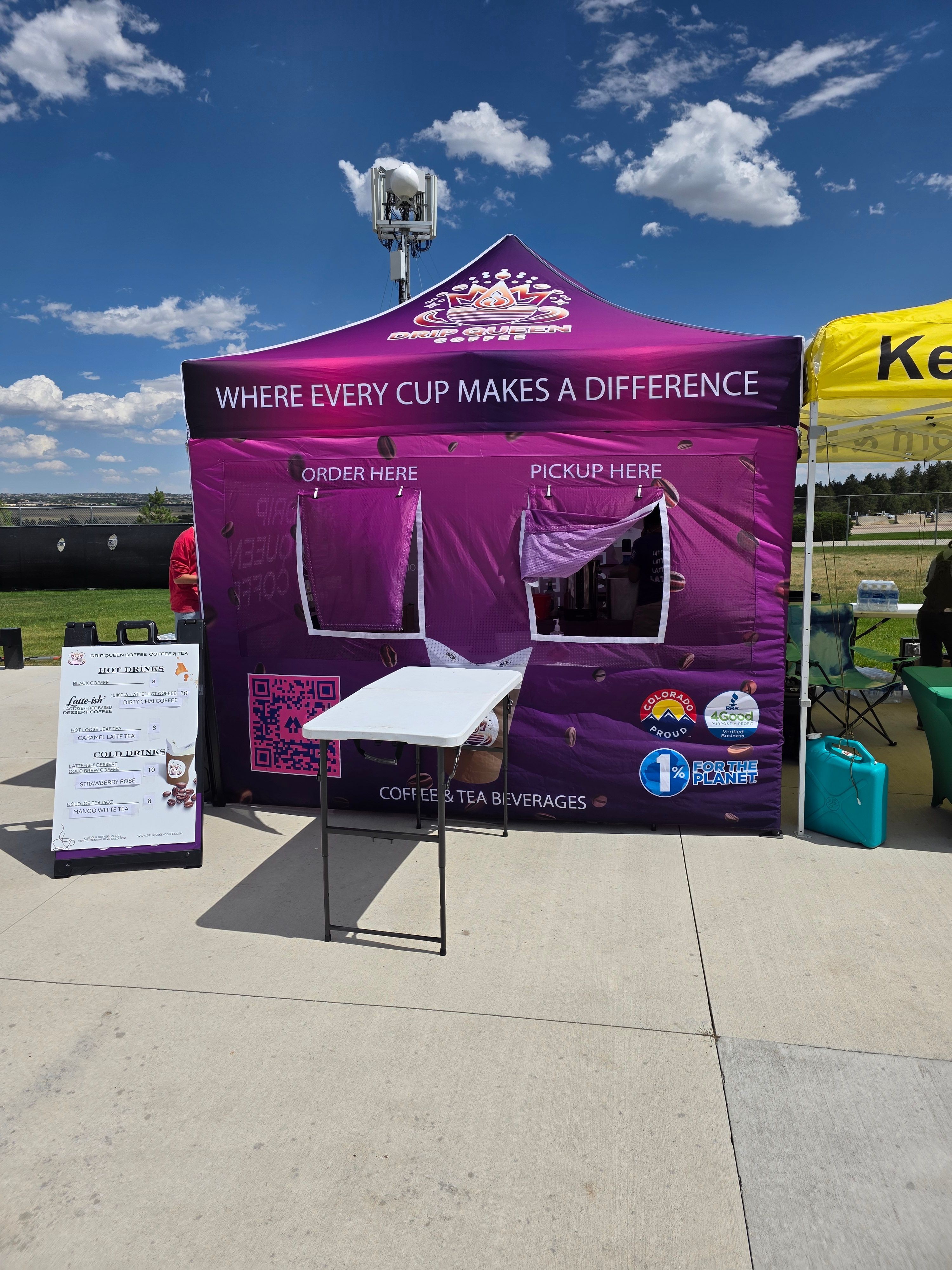 Purple booth with a banner reading 'WHERE EVERY CUP MAKES A DIFFERENCE' and a table with a sign, outdoors on a sunny day.