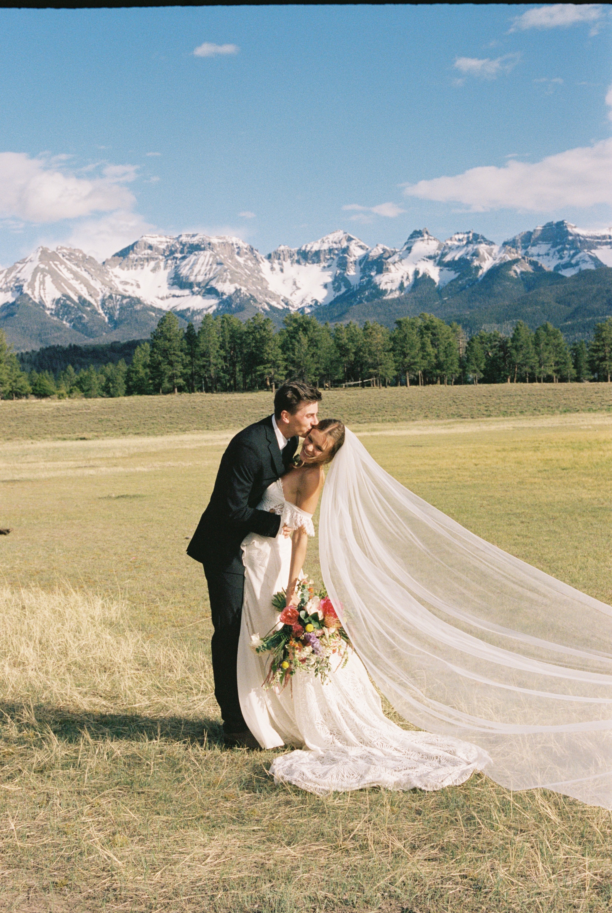 A bride and groom kiss outdoors with mountains and trees in the background, bride holding a bouquet, bride's veil flowing.