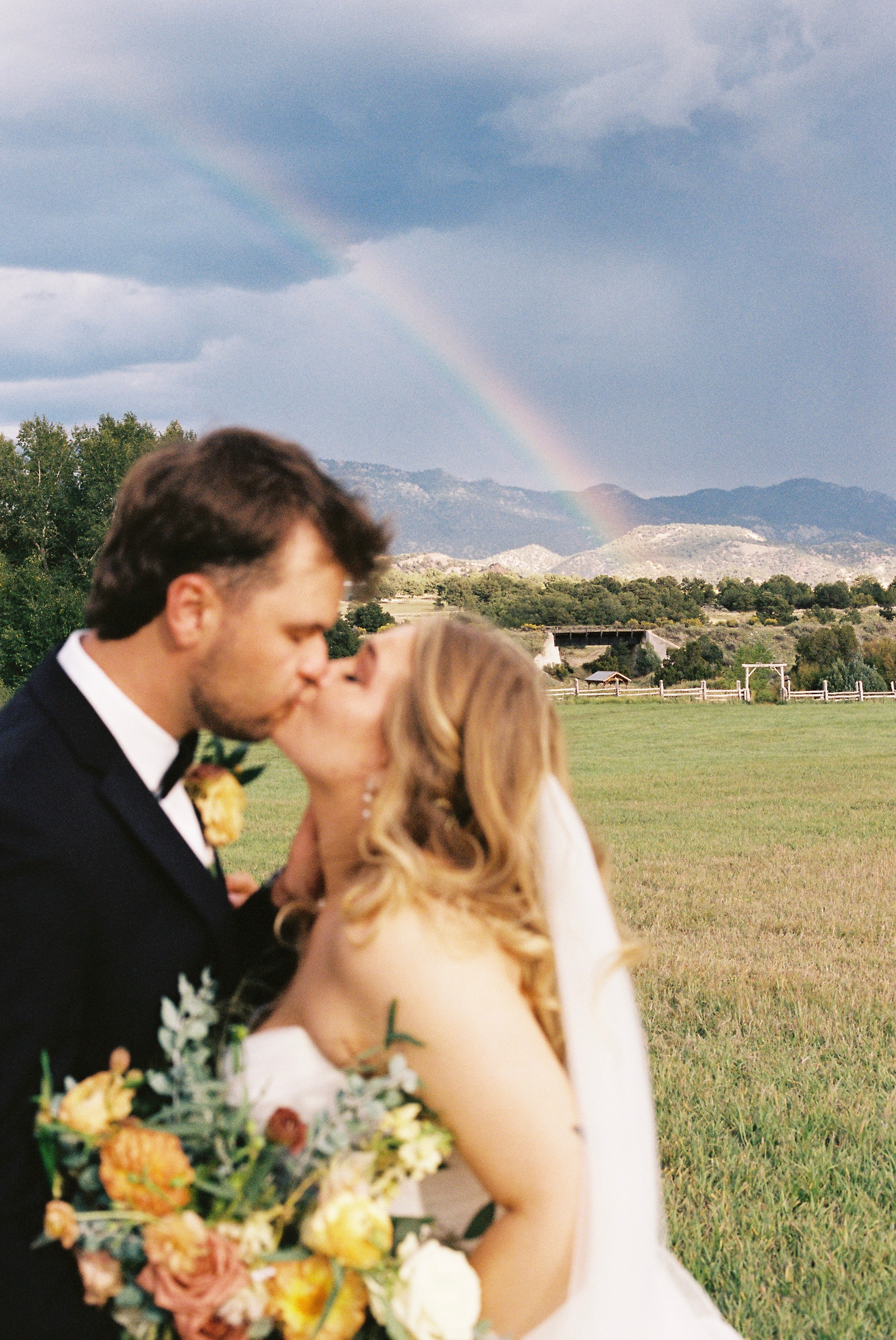 A couple in wedding attire kissing outdoors with a rainbow and mountains in the background.