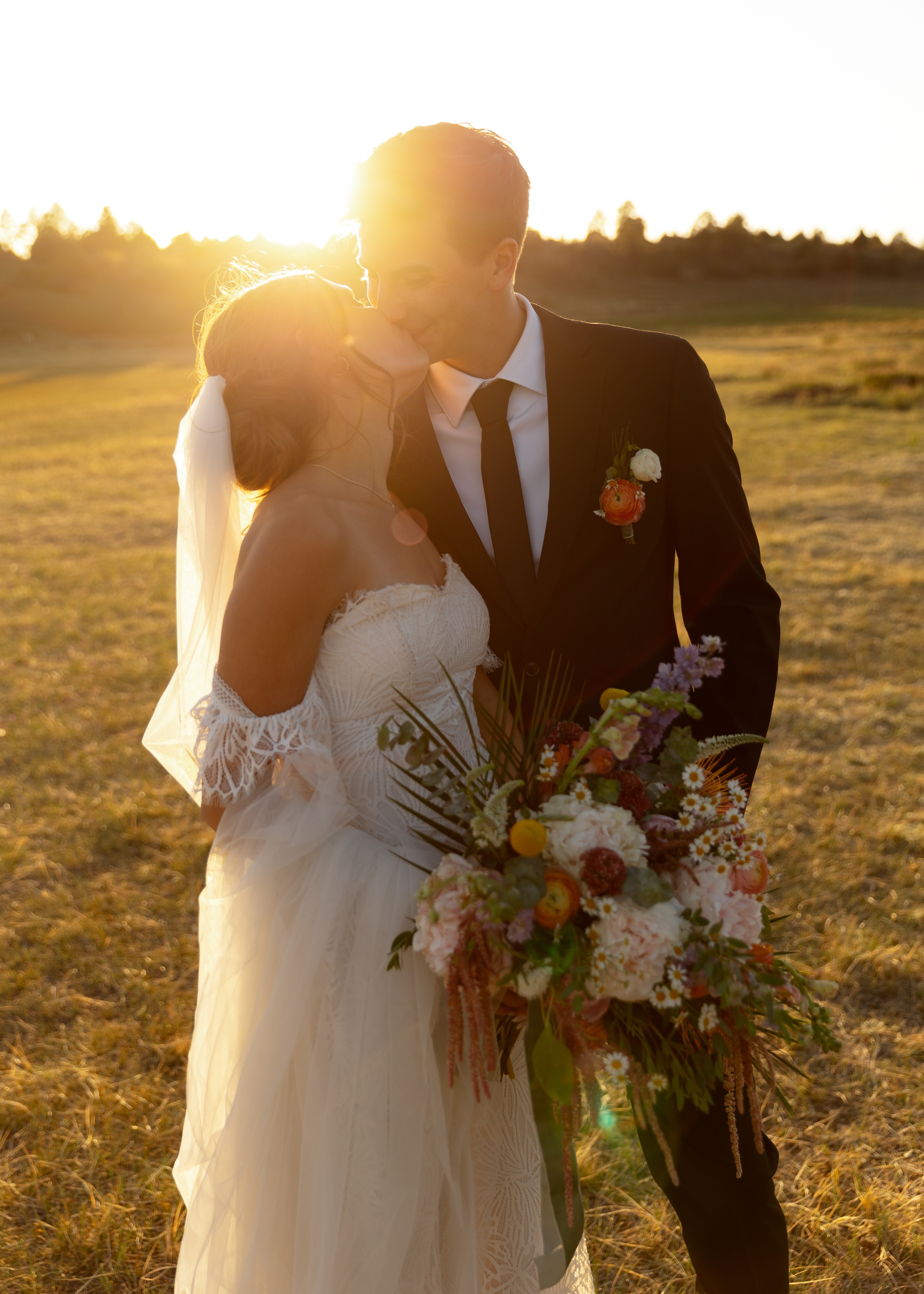 A bride and groom stand outdoors at sunset, with the bride holding a large bouquet of flowers.