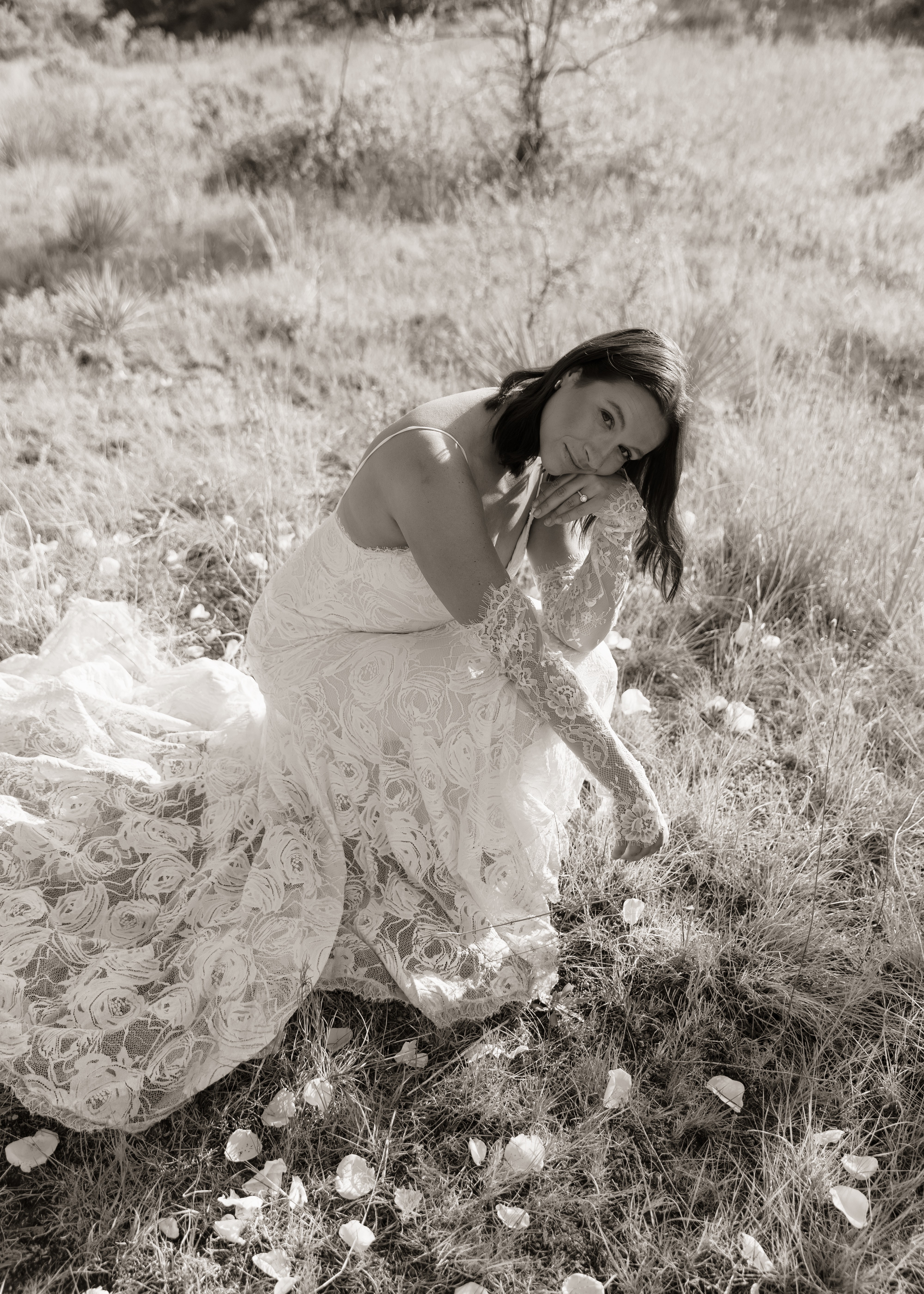 Woman in a long dress sitting on grass in a field, looking down, with trees in the background.