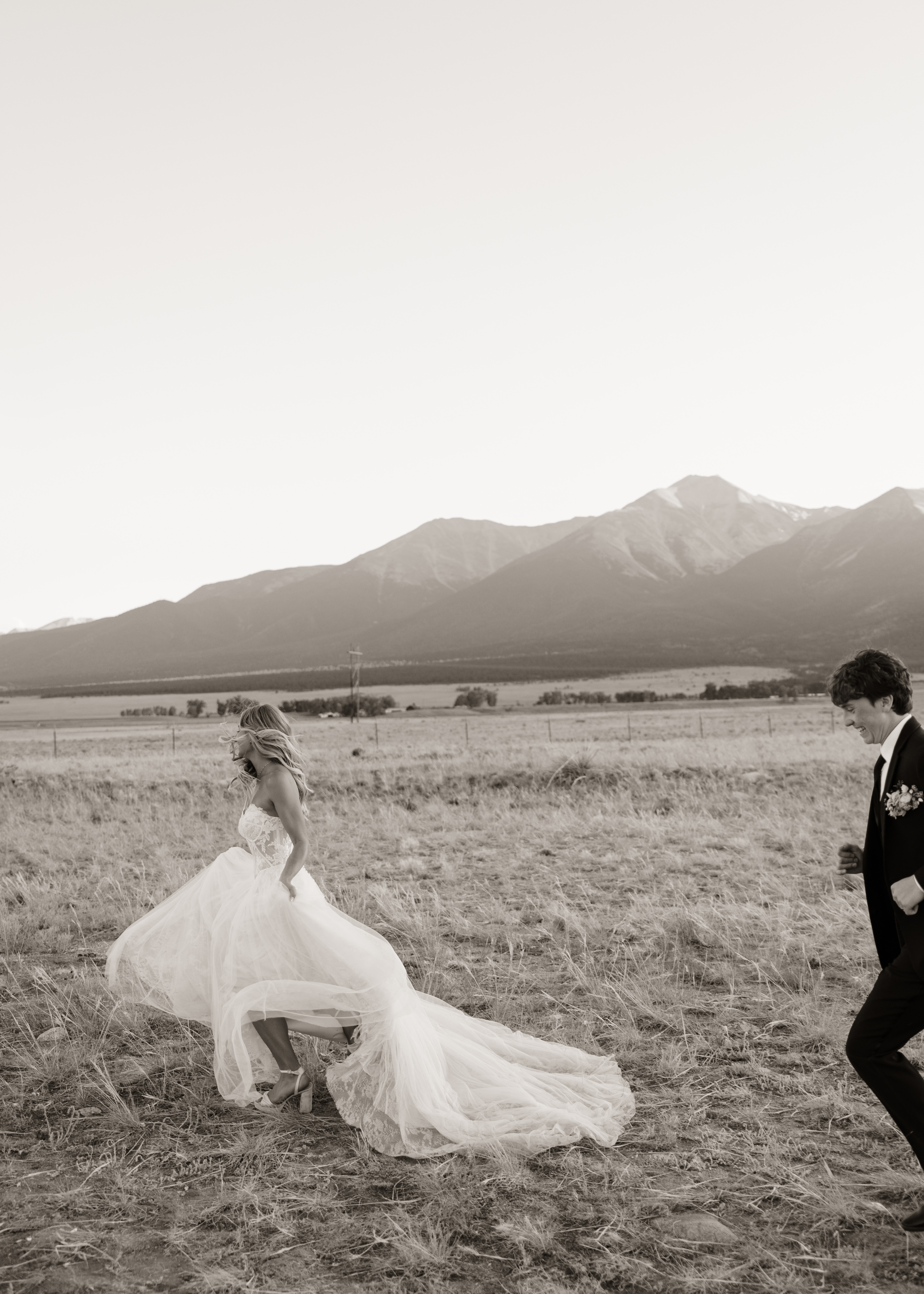 A bride in a wedding dress sitting on the ground in an open field with mountains in the background.