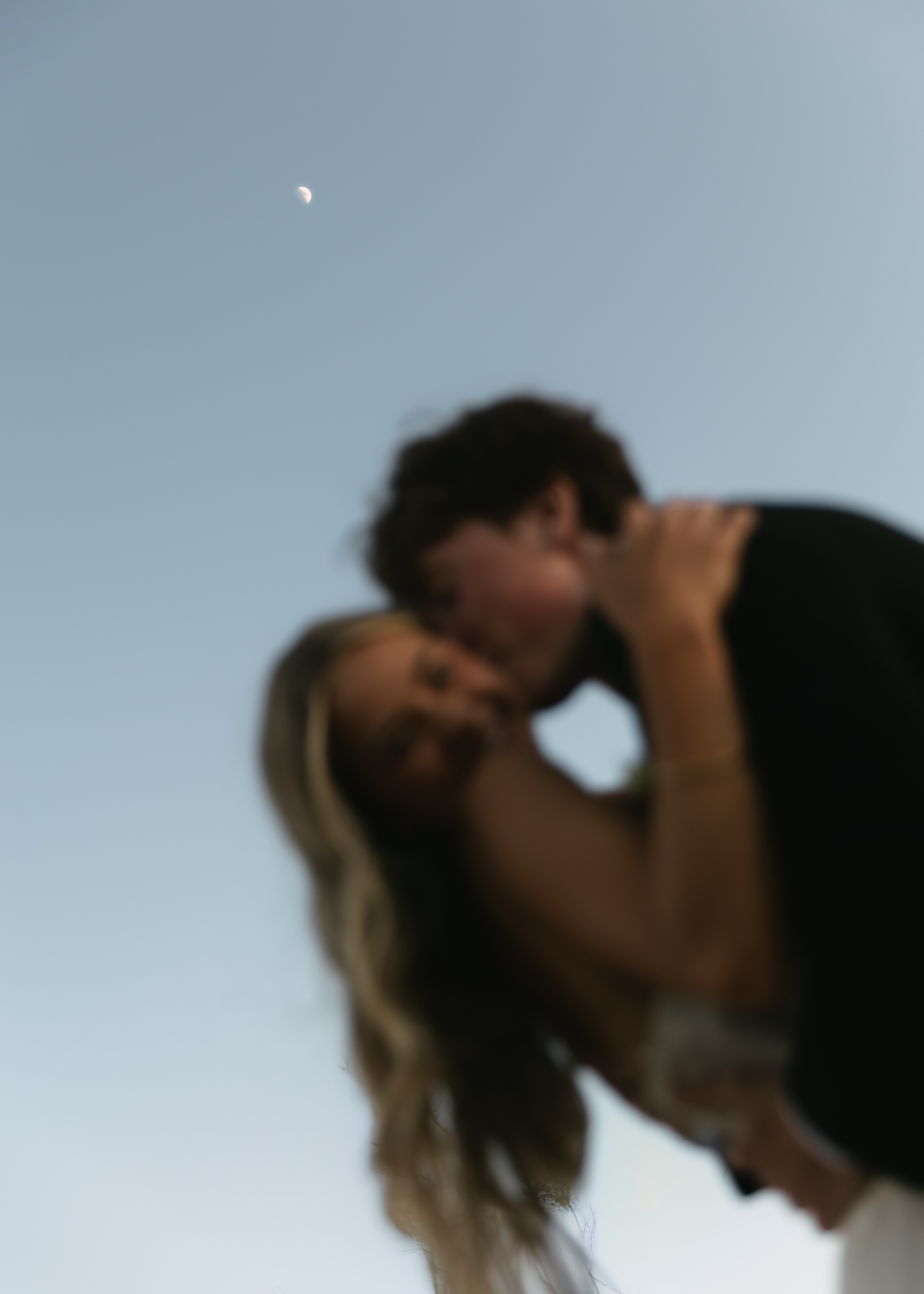 Two people kissing outdoors with a clear sky and visible moon in the background.