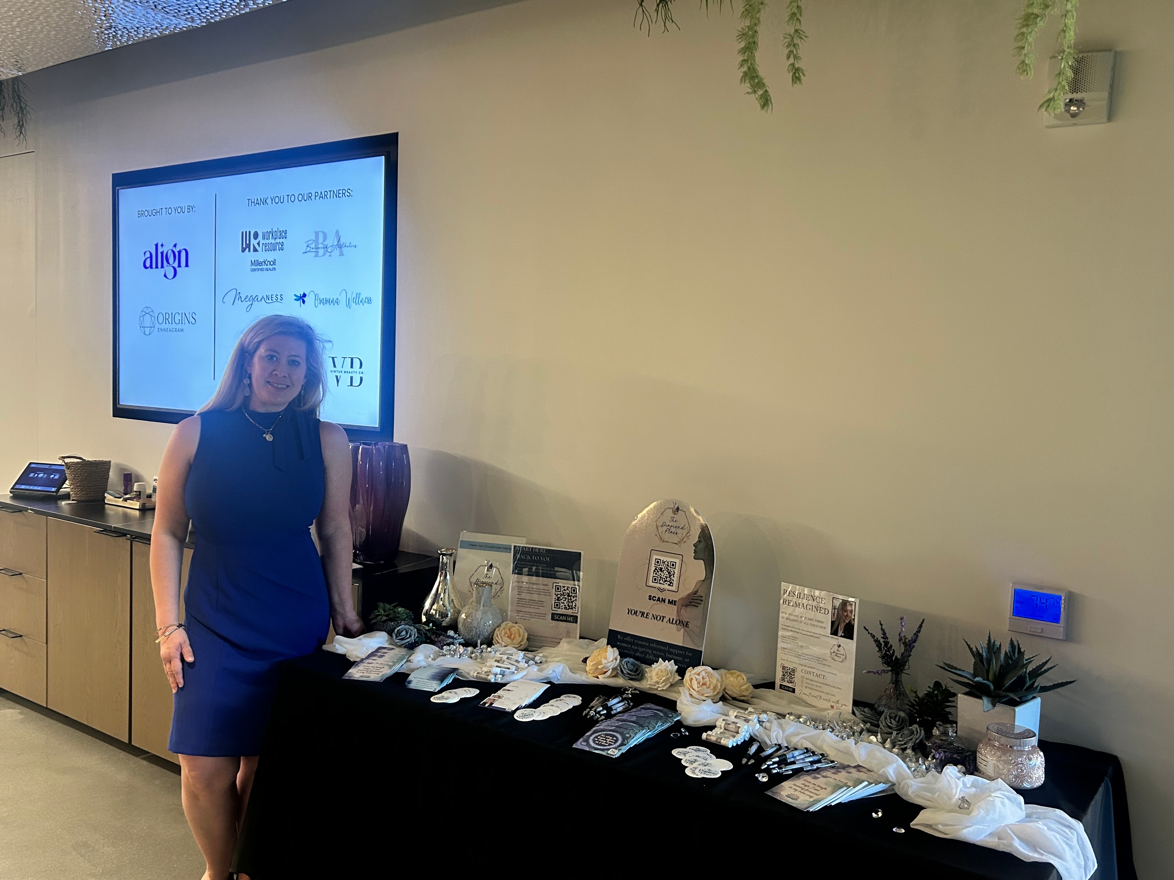Woman in a blue dress standing next to a table with various items, in a room with a large screen on the wall.