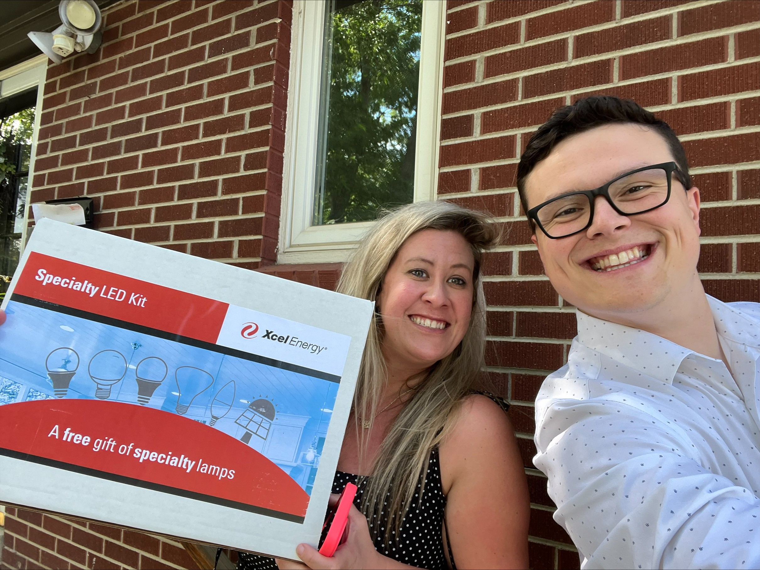 Two people smiling outdoors in front of a brick house, holding a box labeled 'Specialty LED Kit'.