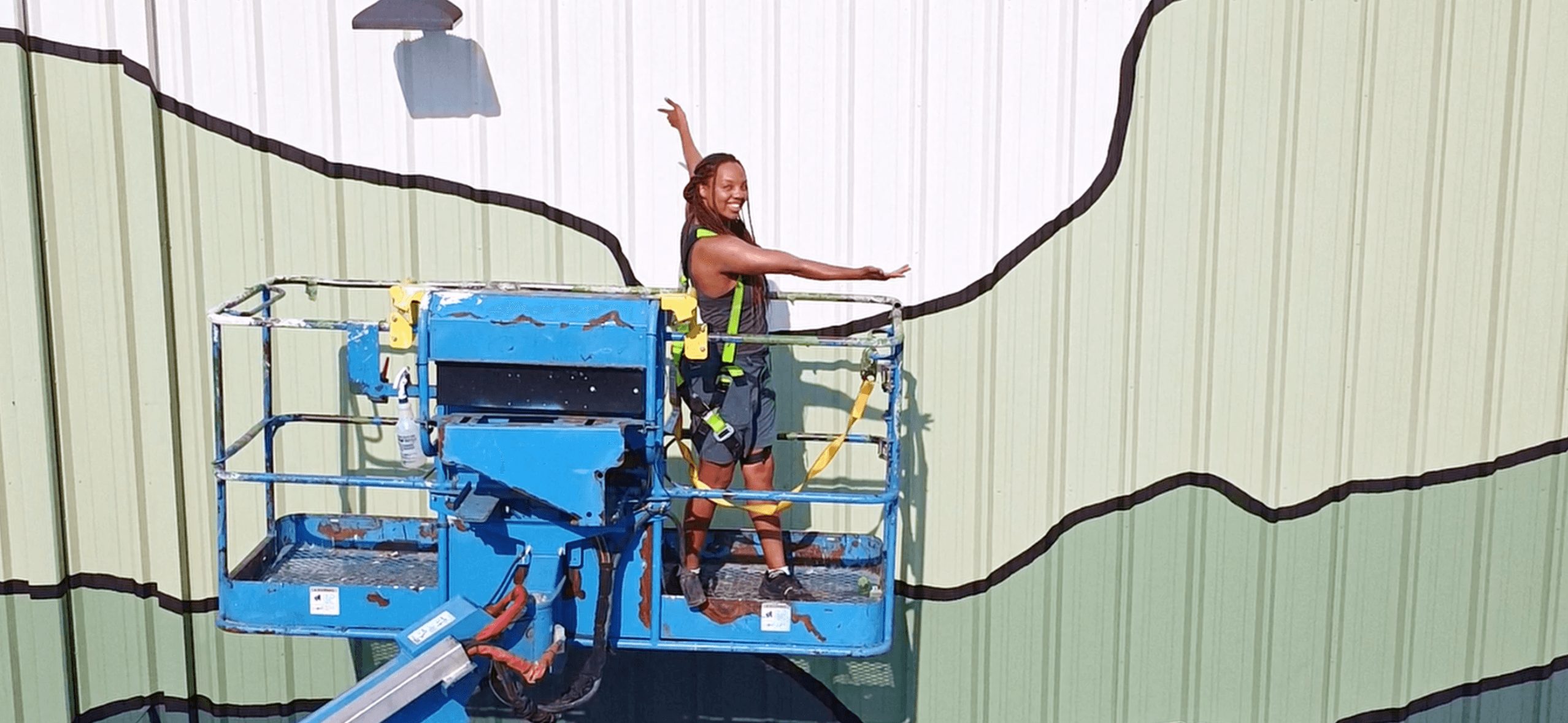 Person on a lift working on a large mural with abstract black lines on a green background.
