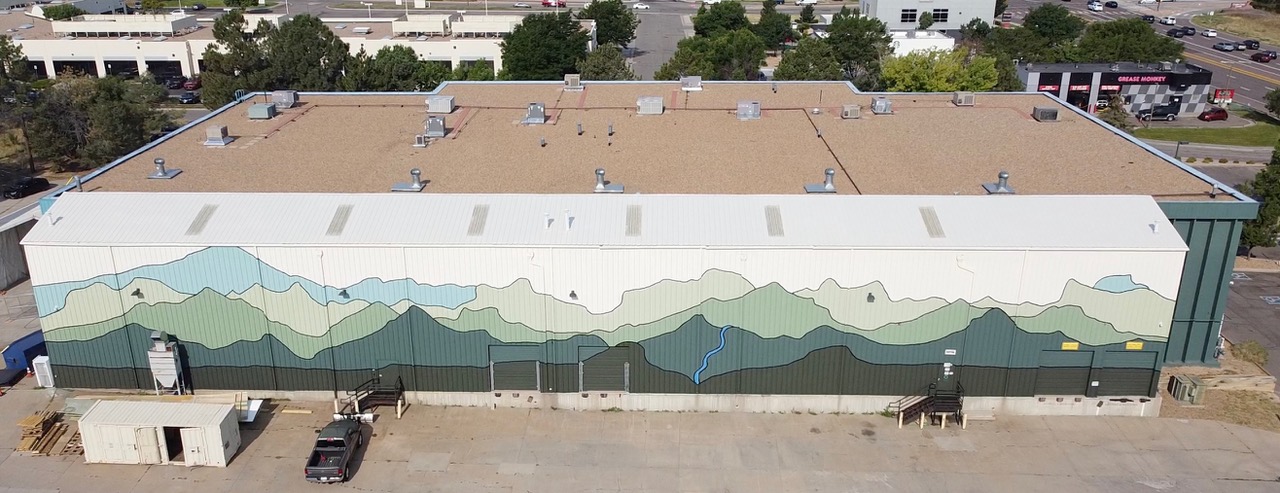 Large mural of mountain landscape painted on building wall, with parking lot and trees in background.