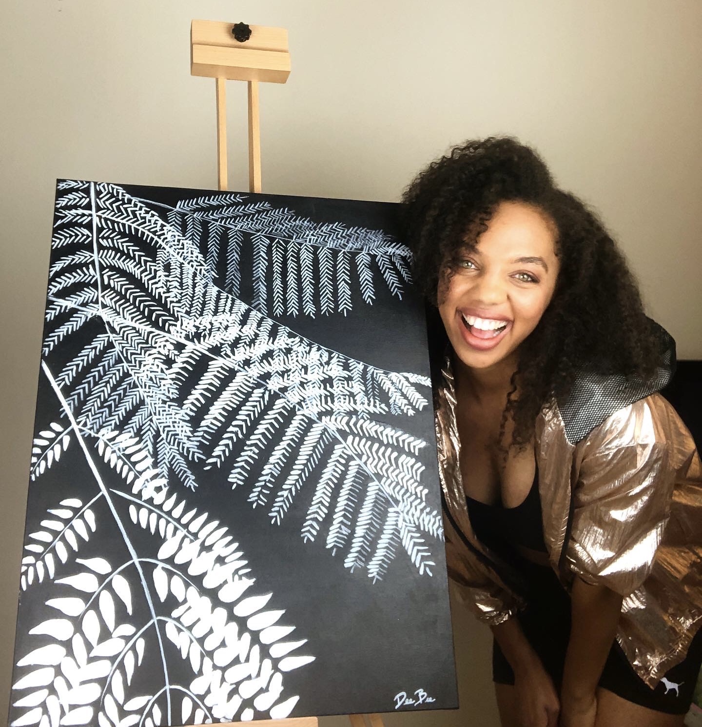 Woman with curly hair smiling and leaning beside a blackboard with white fern leaf drawings, in a room with a beige wall.