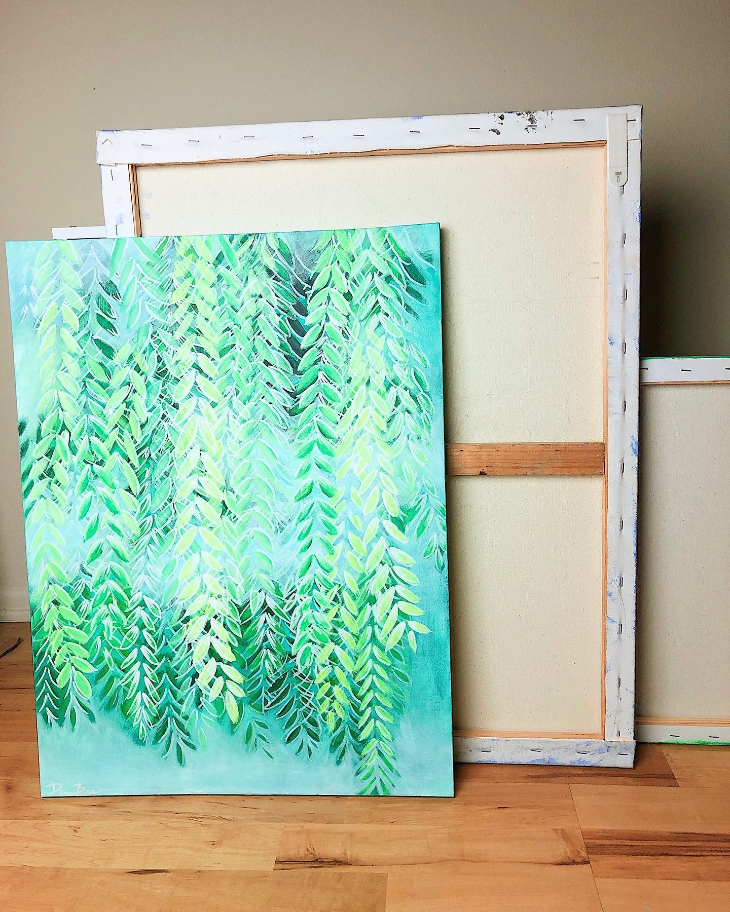 Painting of green hanging leaves leaning against a wooden frame on a hardwood floor.