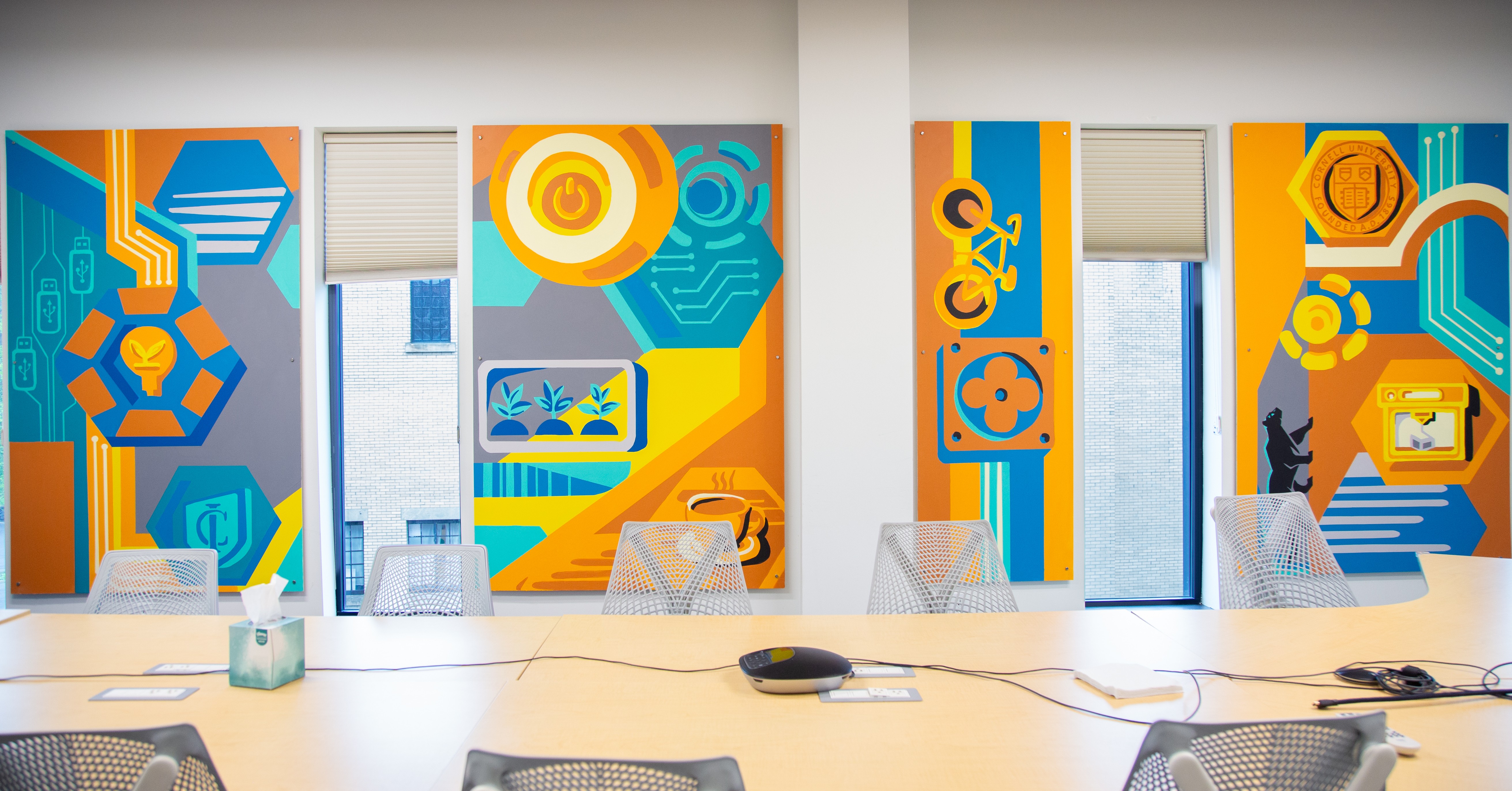 Conference room with colorful abstract wall art, a long table, chairs, and electronic devices, with large windows in the background.