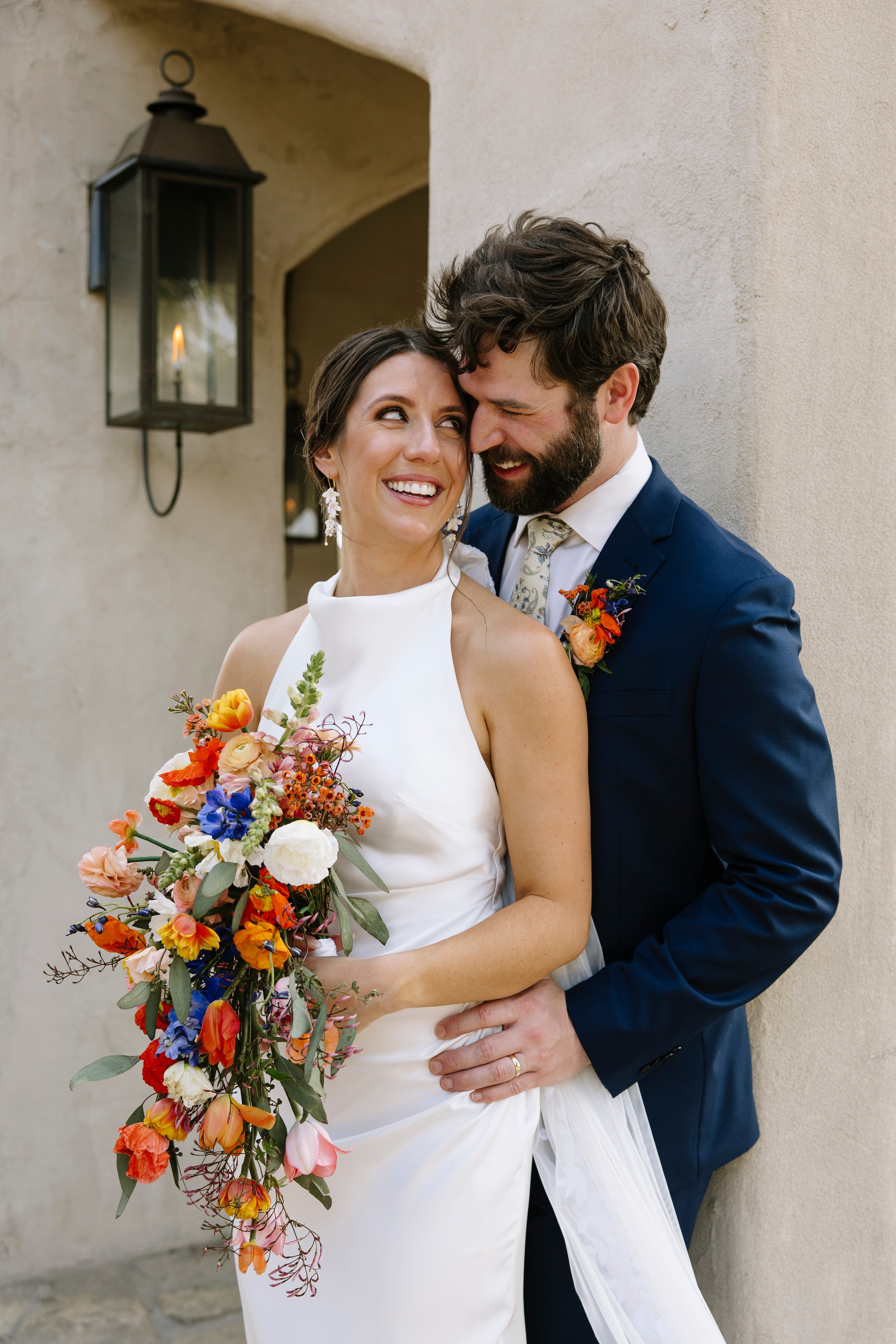 A smiling woman in a white dress holding a colorful bouquet, standing next to a man in a dark suit, outside a building.