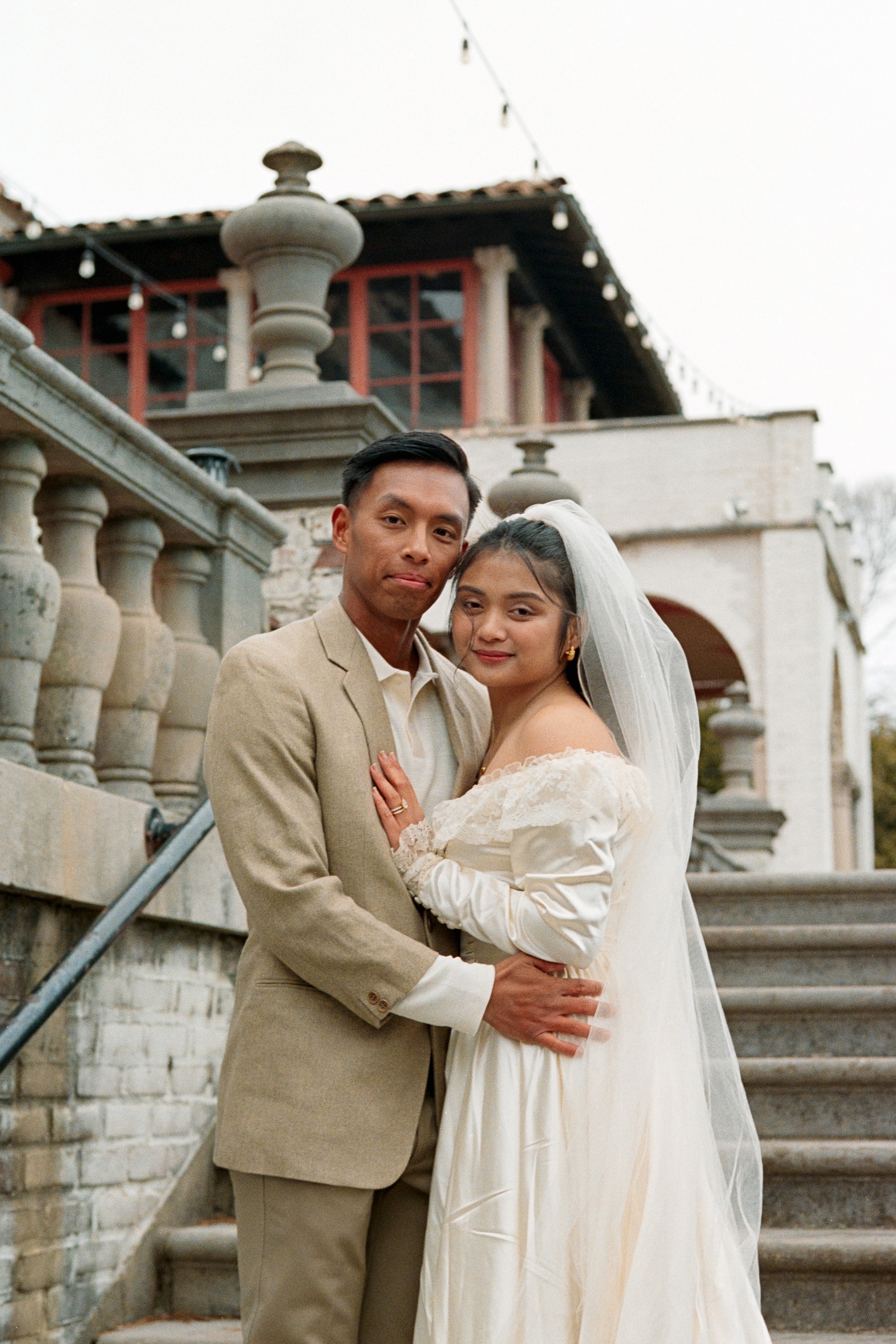 A couple in wedding attire standing on stairs outside a building with traditional architecture, holding each other.