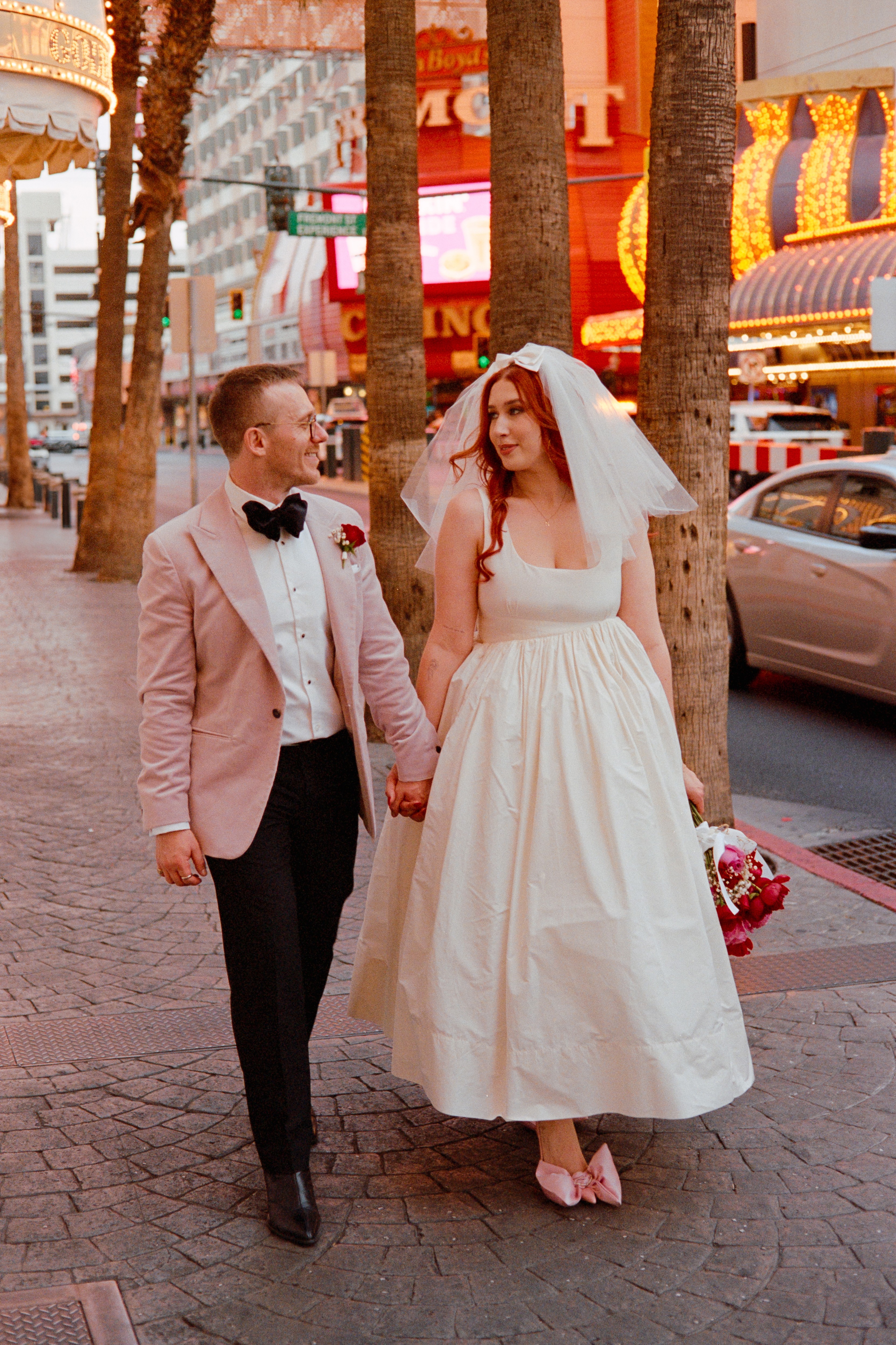 Bride and groom holding hands on city sidewalk, surrounded by trees and buildings, with cars and bright lights in background.