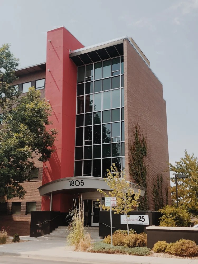 Modern building with glass facade, red and brown exterior, and the number 1805 on the entrance, surrounded by trees and plants.