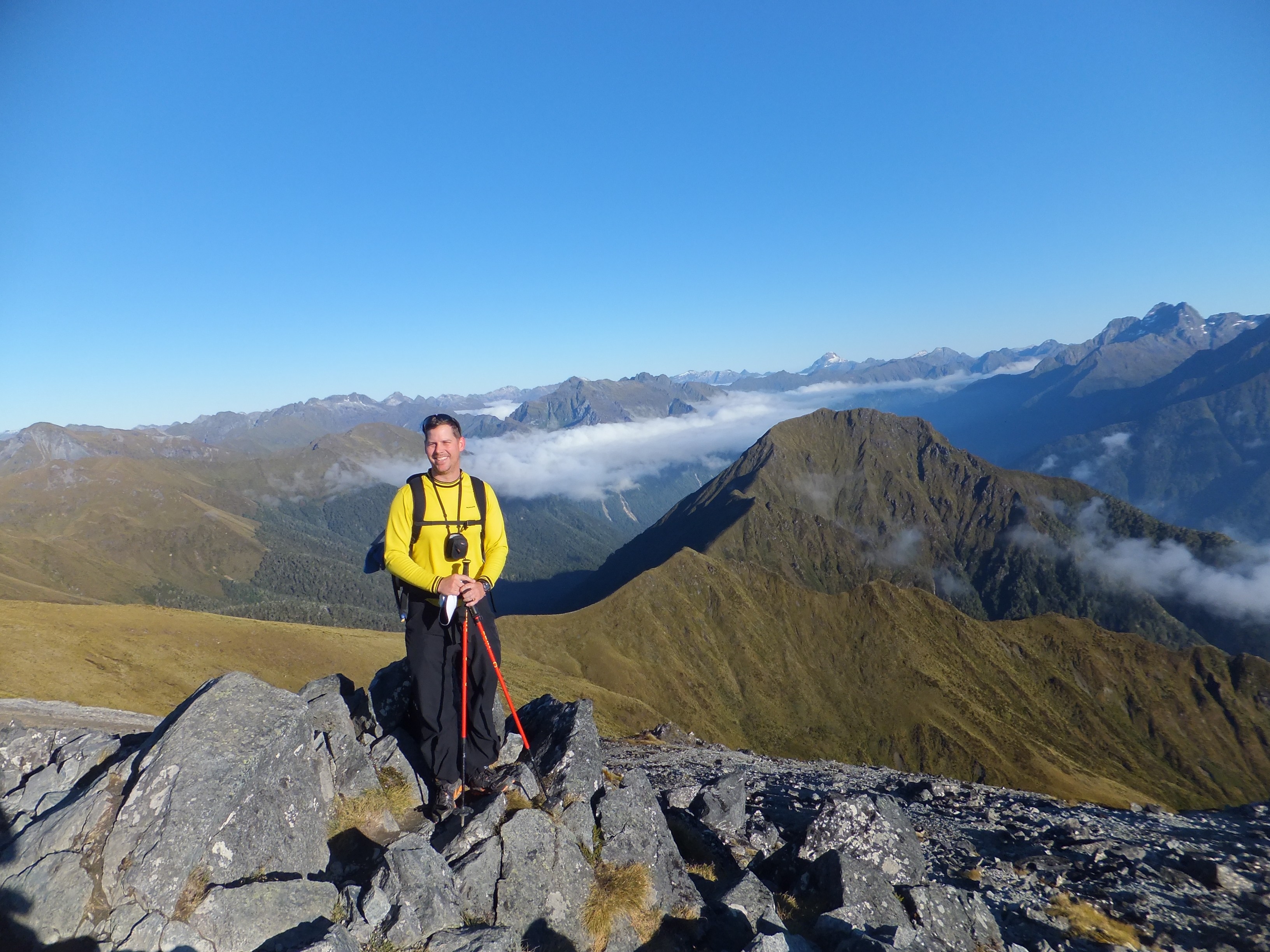 Person standing on rocky mountain terrain with a yellow jacket, mountains and clouds in the background.