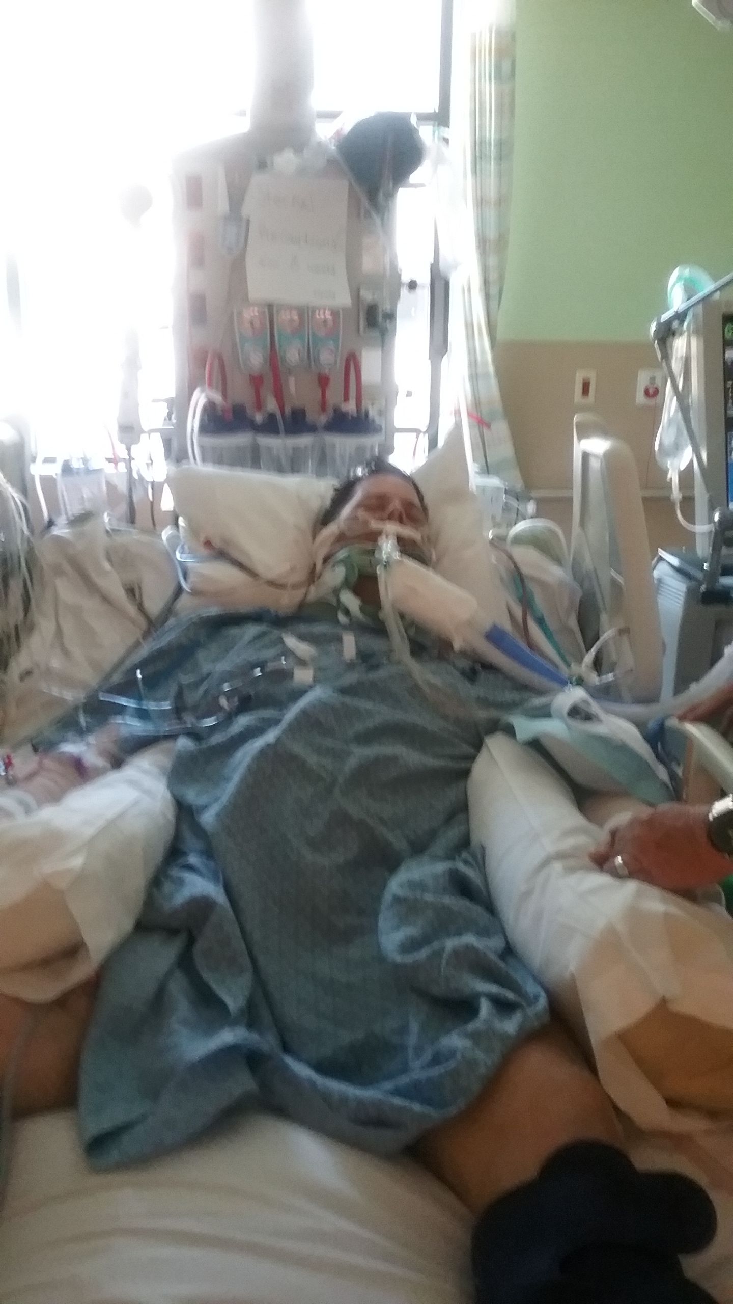 Person lying in hospital bed with medical equipment and healthcare staff nearby, in a clinical setting.