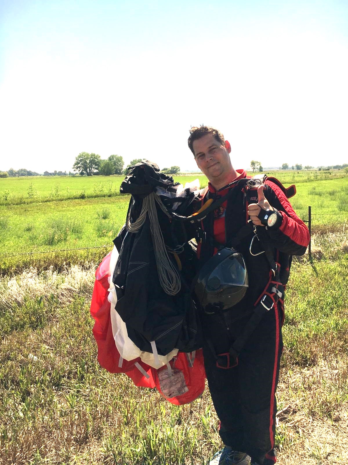Person standing outdoors in a grassy field holding a large backpack and gear, smiling at the camera.