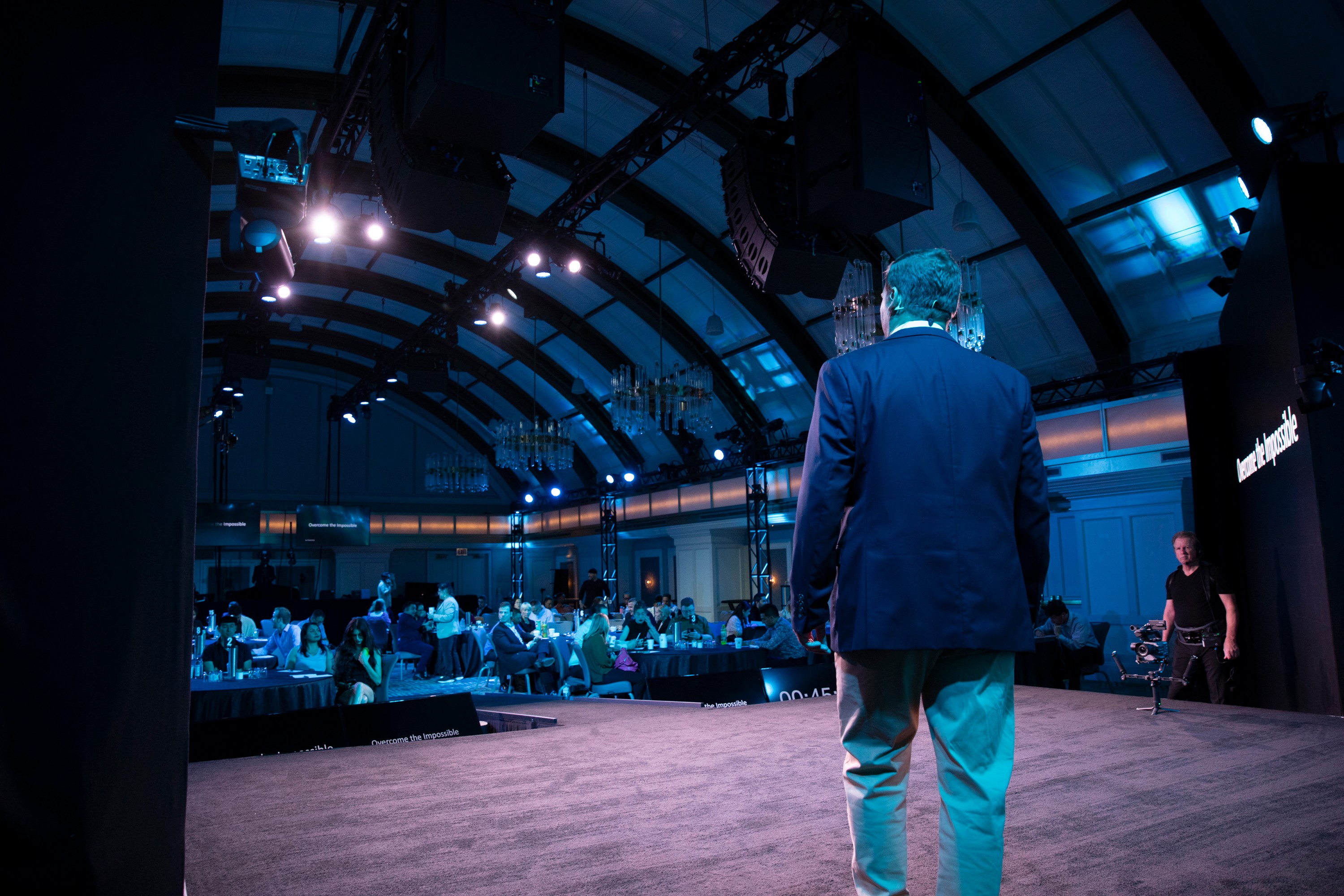 A man in a suit stands on stage facing an audience in a large, curved venue with blue lighting.