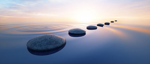 Seven stones arranged in a line on calm water, with a sunset or sunrise in the background.