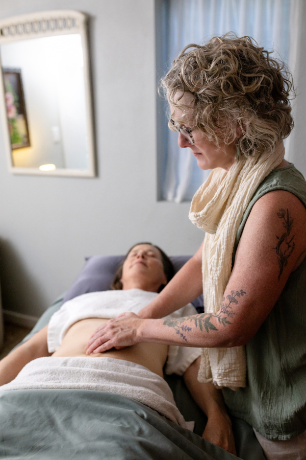 A woman with curly hair and tattoos leans over a person lying in bed, in a room with a mirror and curtains.