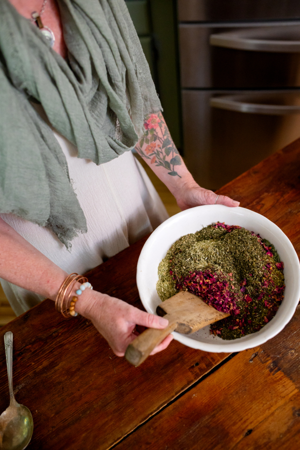 Person holding a bowl of green and purple food, wearing a green scarf and bracelets, standing at a wooden table.