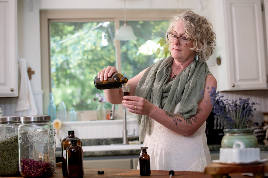 Woman with curly gray hair pouring liquid from a bottle into a small container in a kitchen with a window and plants behind her.