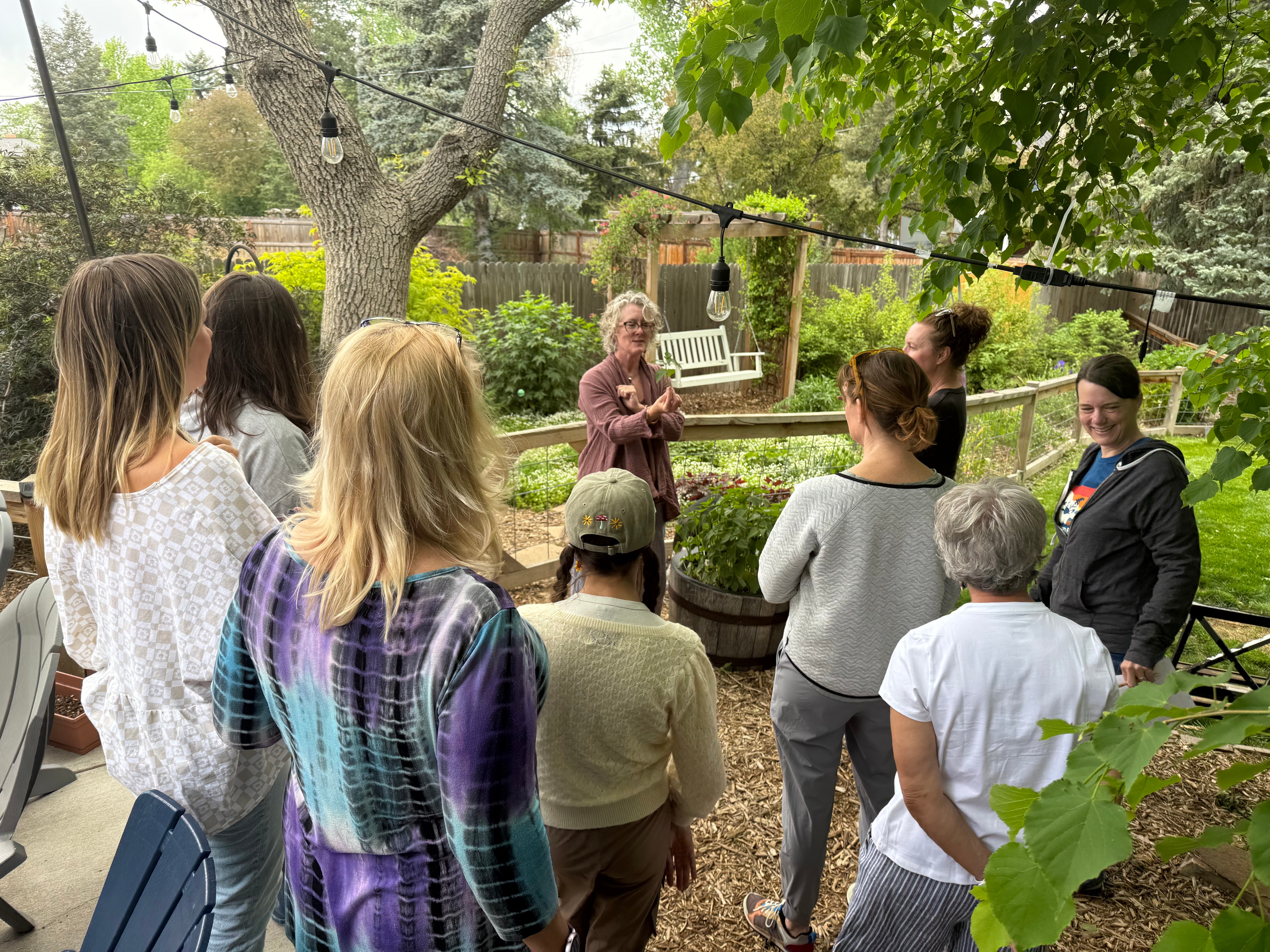 Group of people gathered outdoors around a woman speaking, trees and greenery in the background.