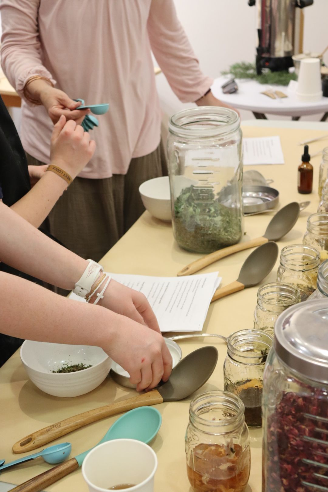 People's hands and arms reaching over a table with jars, spoons, and papers, in a room with a white wall and furniture.