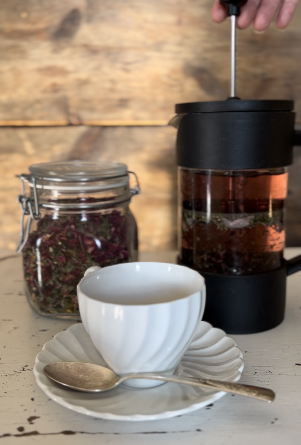 A French press, a jar of dried herbs, and a cup with a saucer and spoon on a wooden surface.
