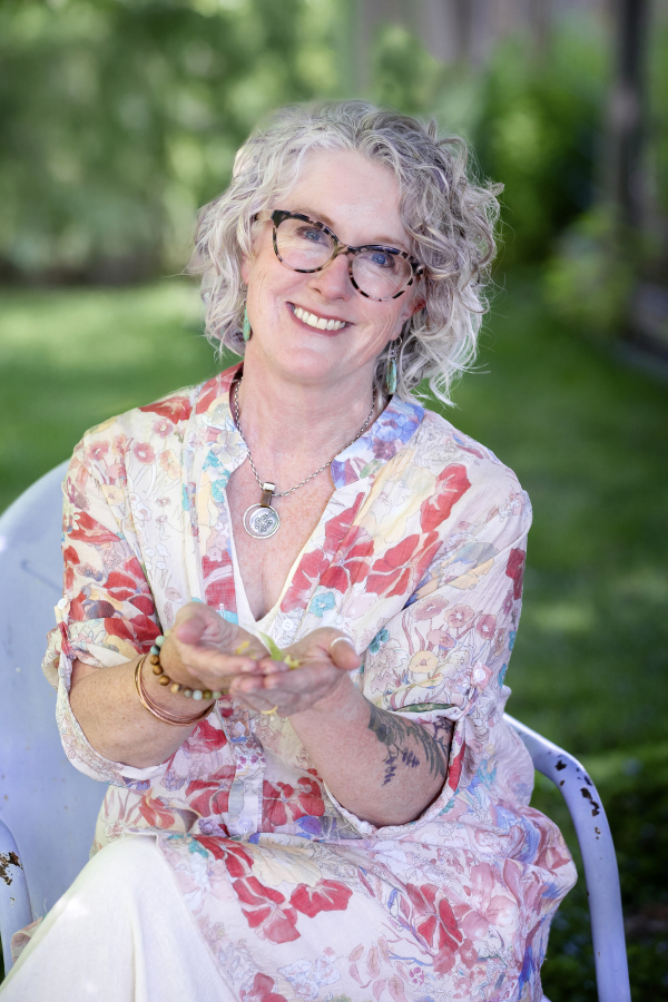 Smiling woman with curly gray hair, glasses, wearing a floral dress, sitting outdoors with greenery in background.