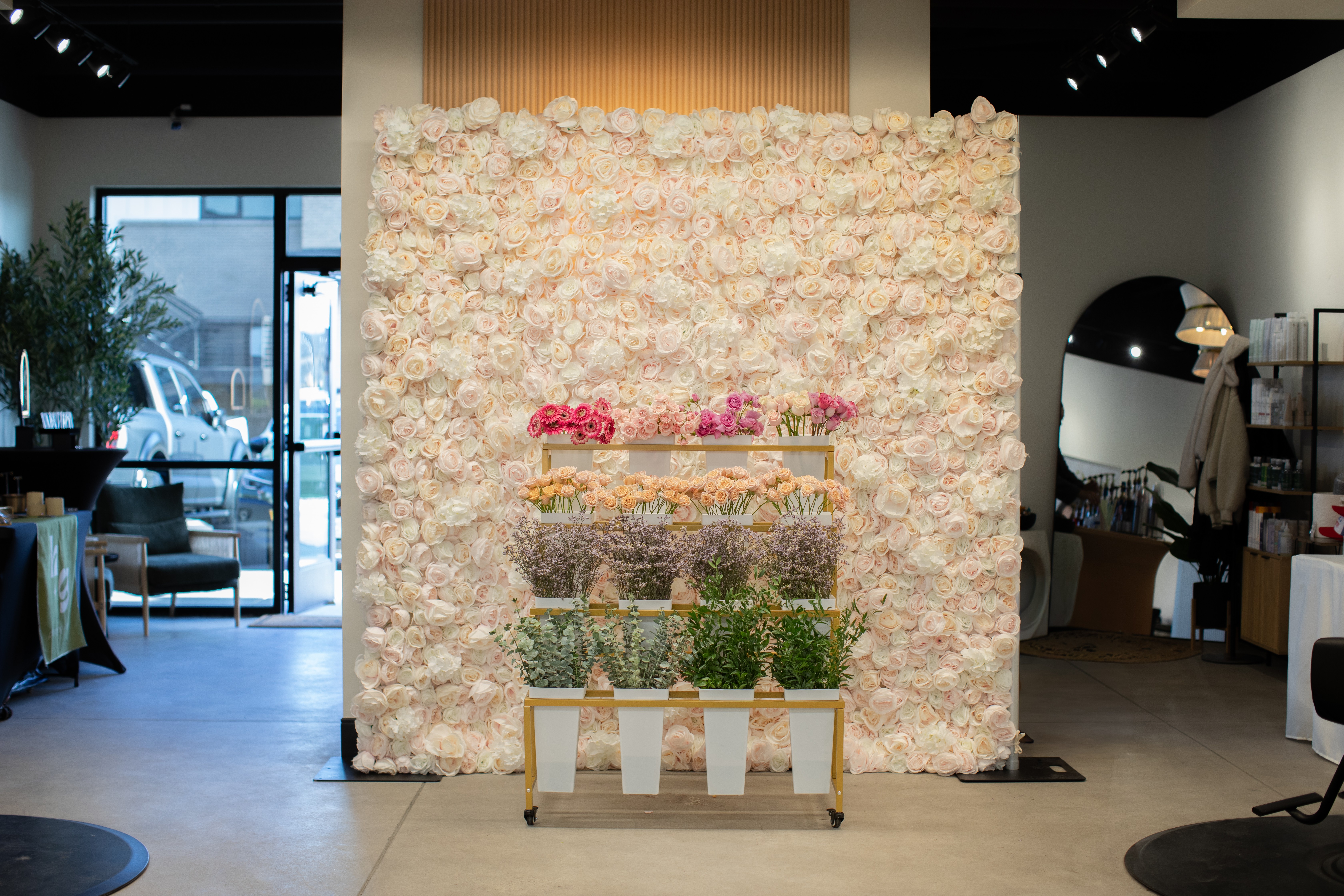 Flower wall with pink, white, and yellow flowers, and potted plants on a stand in a decorated indoor space.