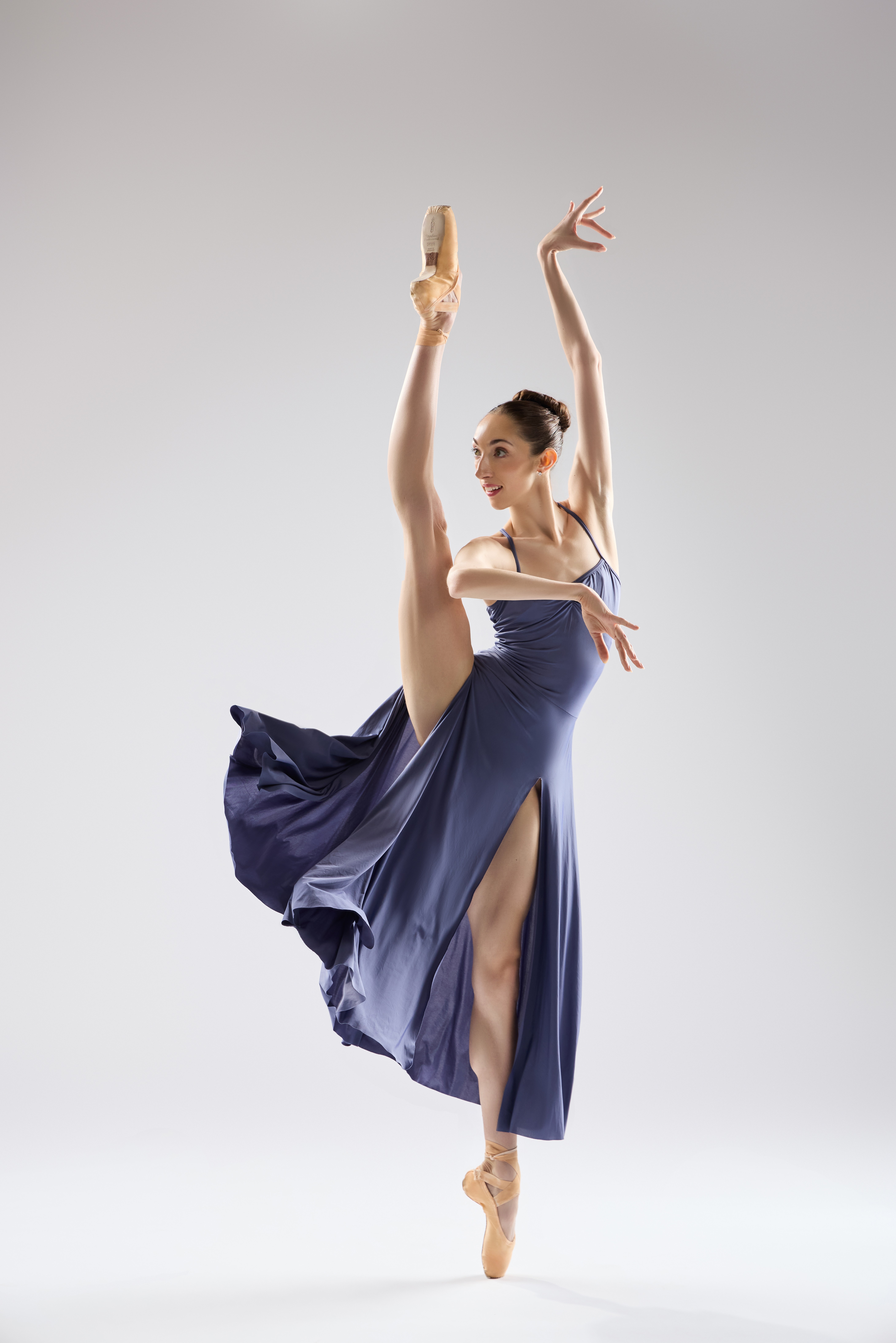 Ballet dancer in a blue dress performs a high leg extension with arms raised, on pointe, against a plain background.