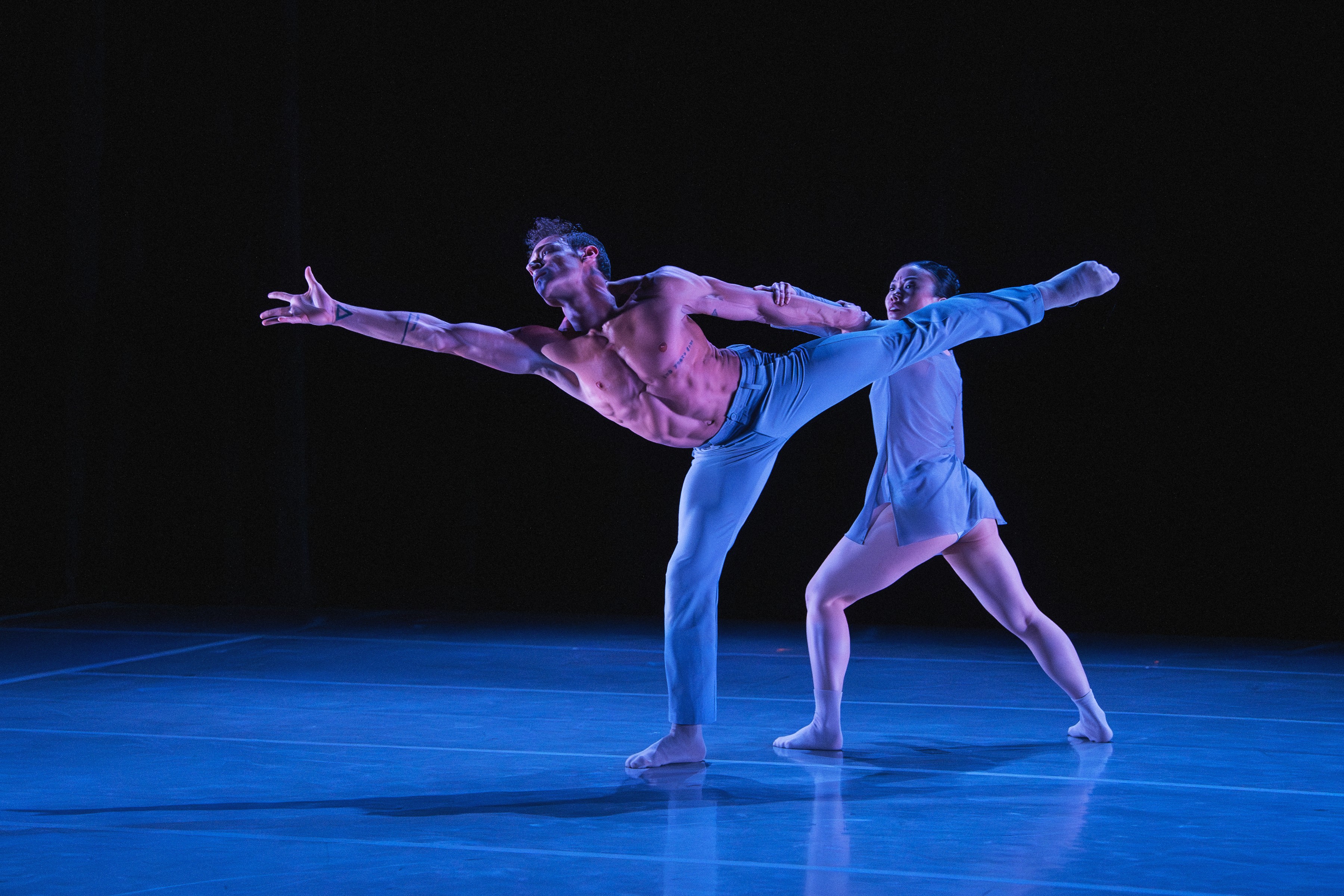Two ballet dancers perform on stage, one with arms extended and the other supporting, against a dark background.
