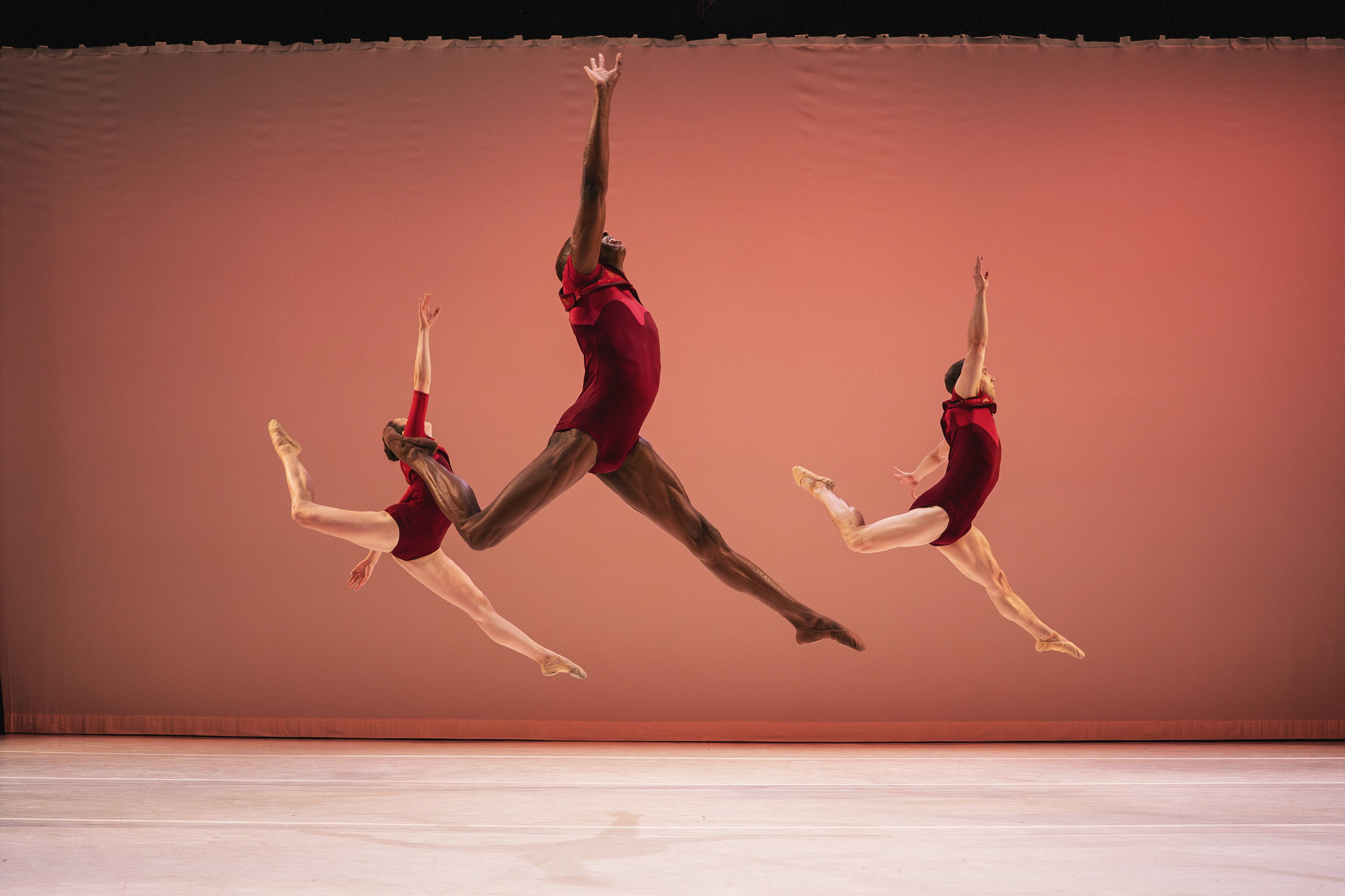 Three dancers in red costumes perform jumps against a pink background, with one in the center and two smaller ones on sides.