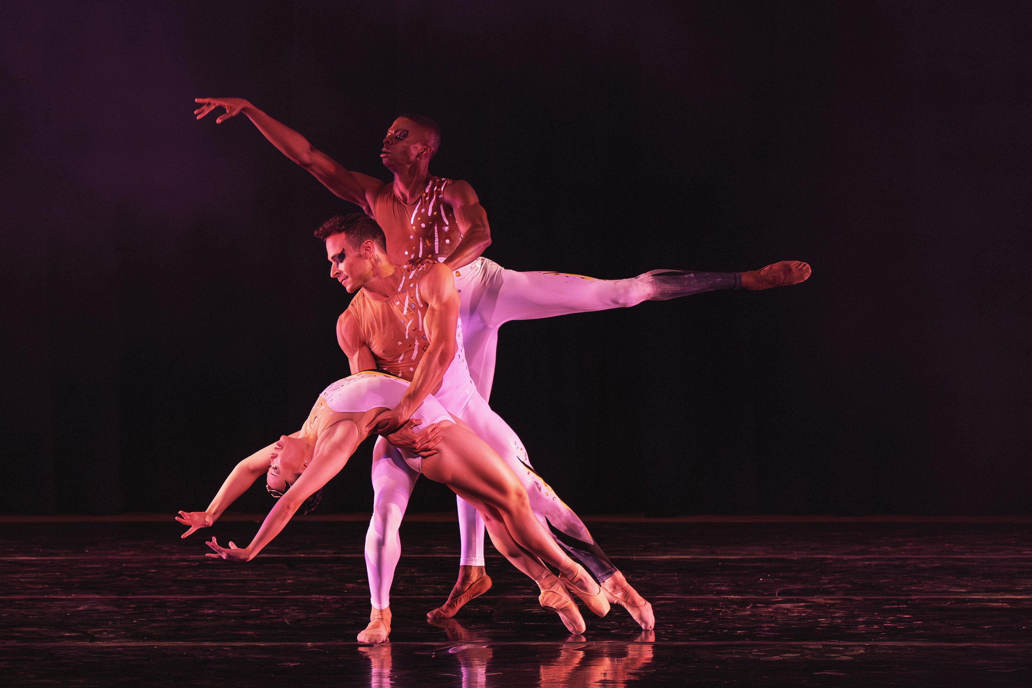 Three ballet dancers perform on stage, with one dancer in a backbend and others supporting and extending limbs.