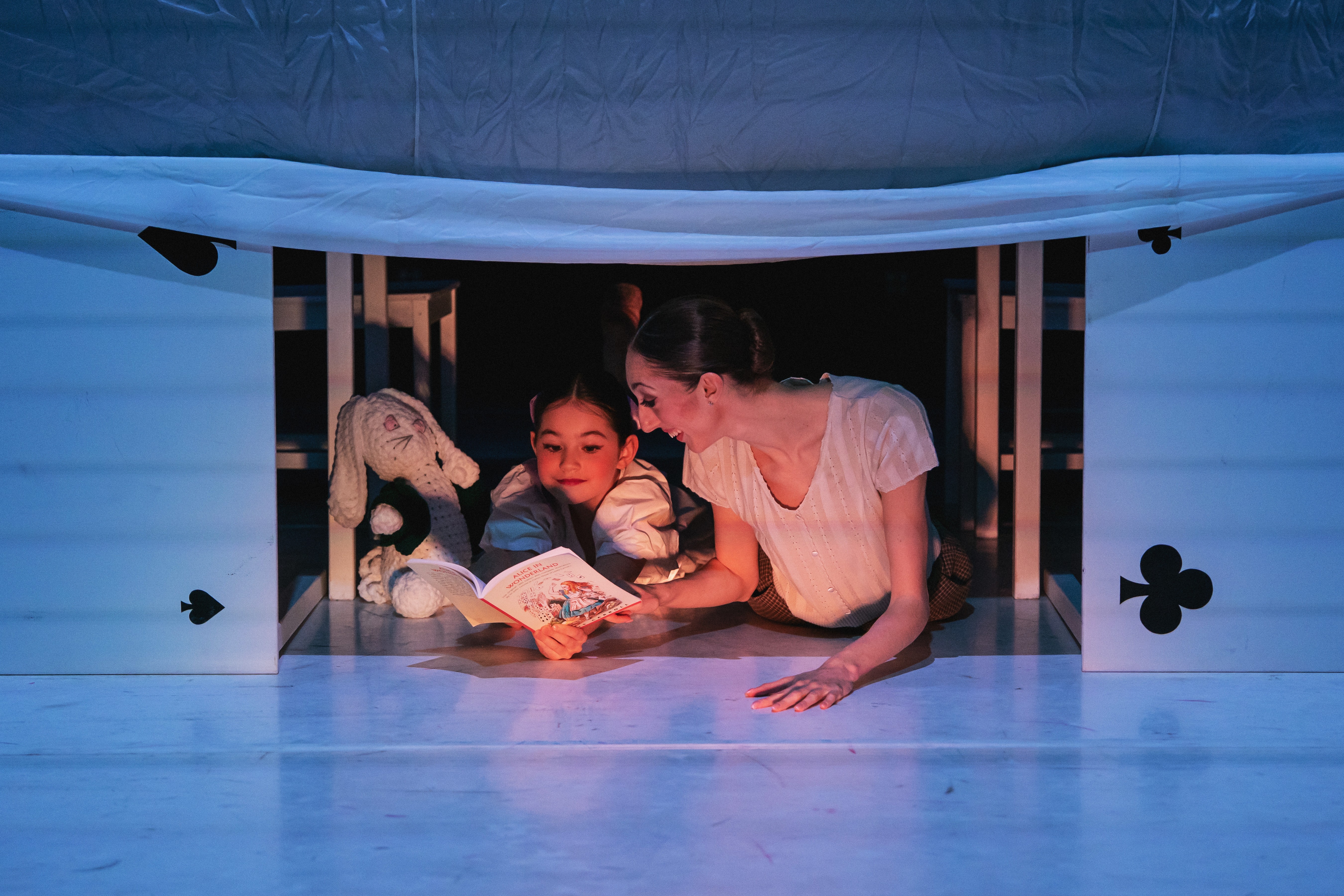 Two people, a child and an adult, reading a book together inside a playhouse with a window opening, decorated with clover symbols.