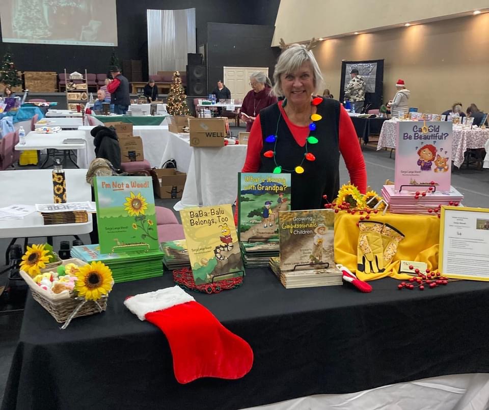 Woman standing behind a table with books, sunflowers, a Christmas stocking, and festive decorations at an indoor event.